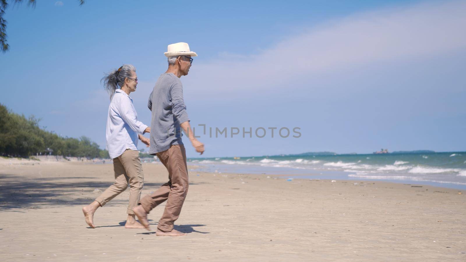 Happy Asian senior man and woman couple holding hands walking to the beach sunny with bright blue sky, Romantic elderly enjoy Travel summer vacation, plan life insurance at retirement couple concept