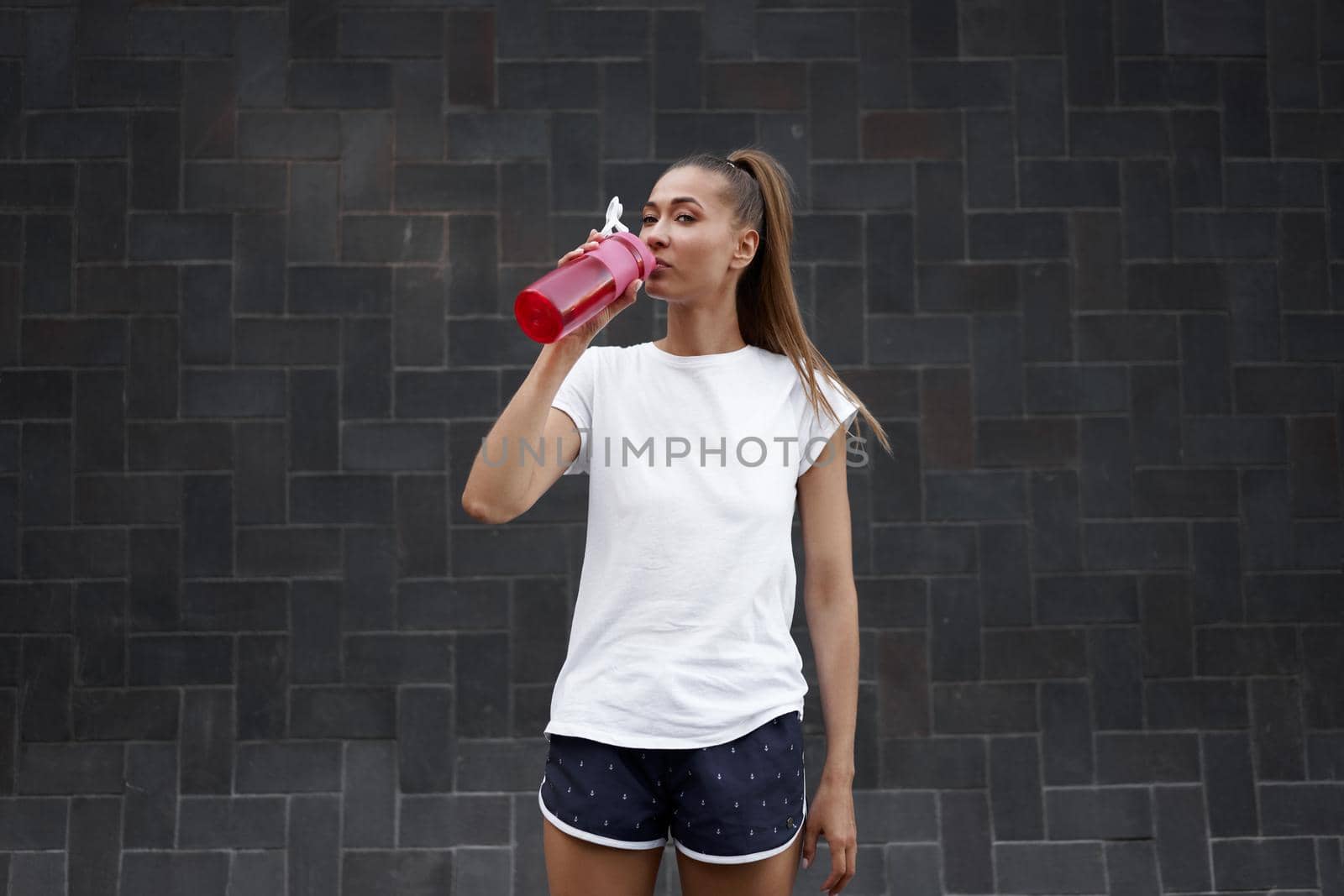 Tired Woman drink water red bottle after morning workout. Young athletic female standing city street black wall background after jogging Healthy lifestyle concept