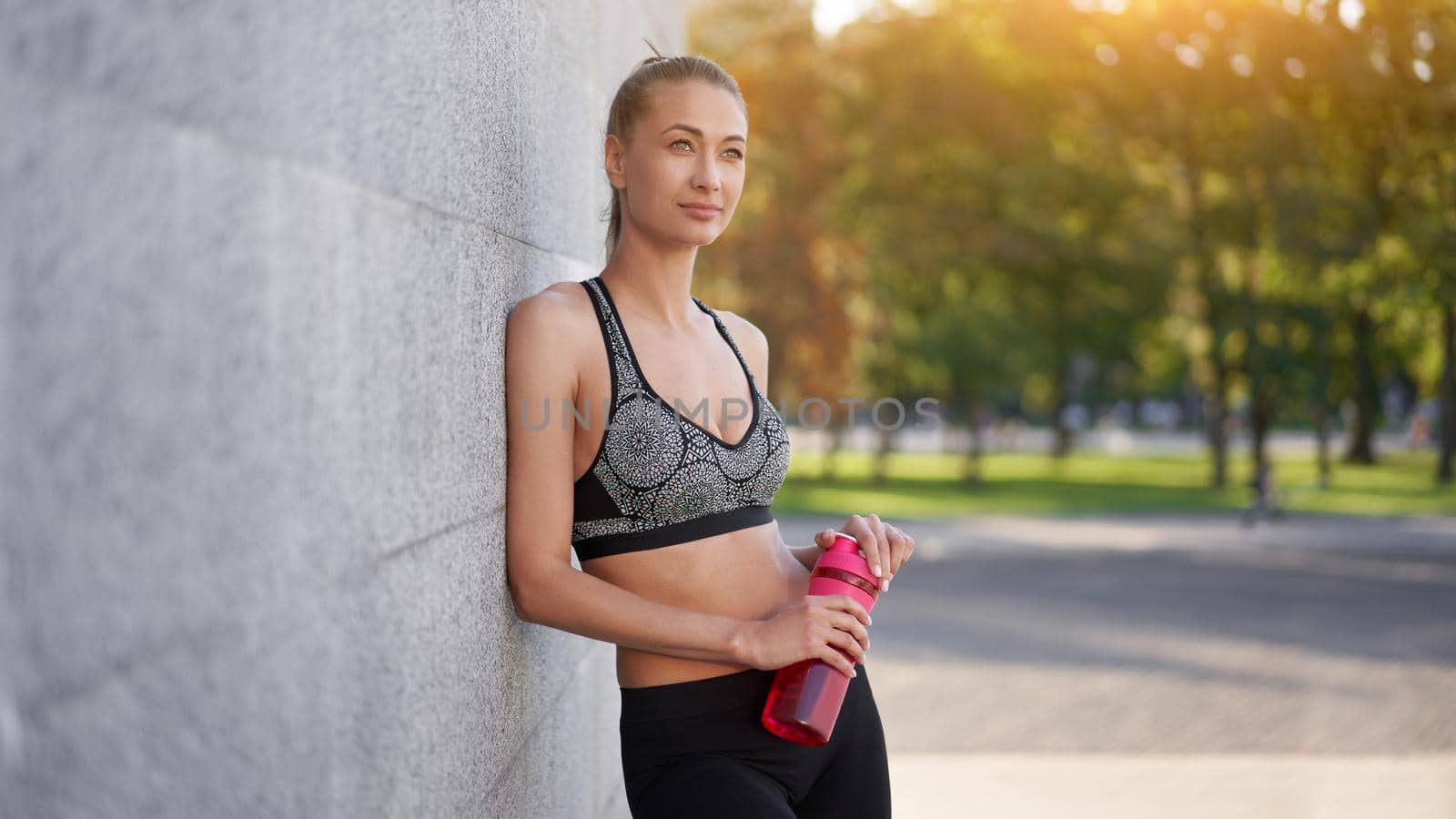 Tired Woman drink water red bottle after morning workout Young athletic female rear view standing leaning wall city street park background after jogging Healthy lifestyle concept Copy space