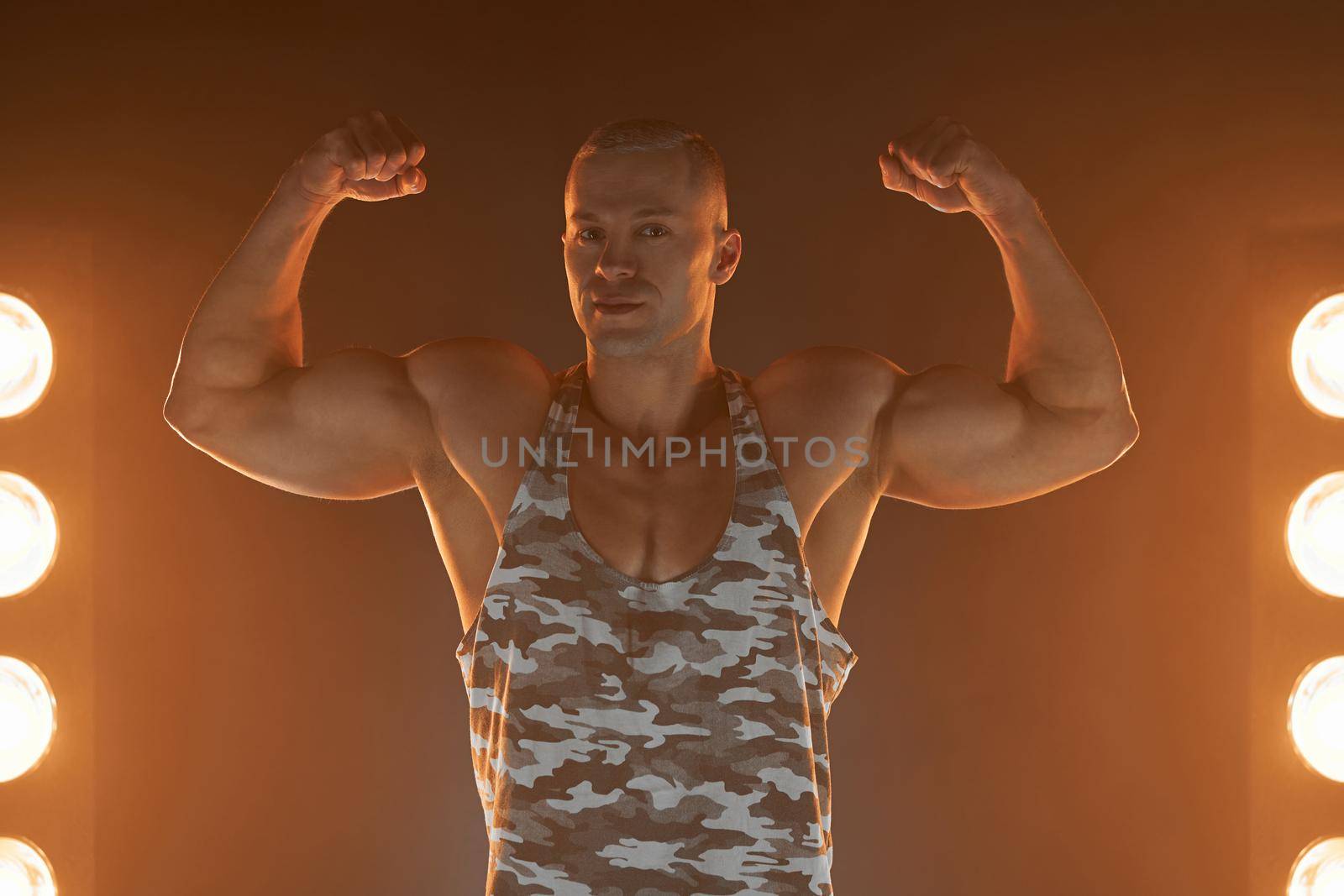 Active lifestyle concept. Professional bodybuilder showing biceps and smiling at camera, lamps illumination and smoke on background