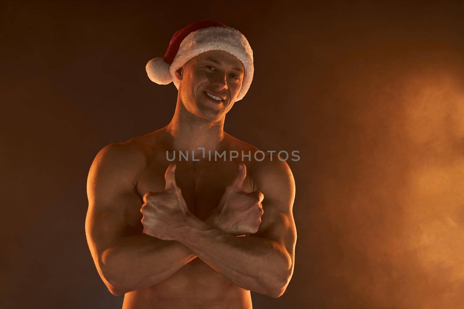 Portrait of muscular man wearing Christmas Santa hat, folded hands and smile on smoky background Macho Shirtless Naked torso stripper Hot sexy Santa guy Seductive male for holiday