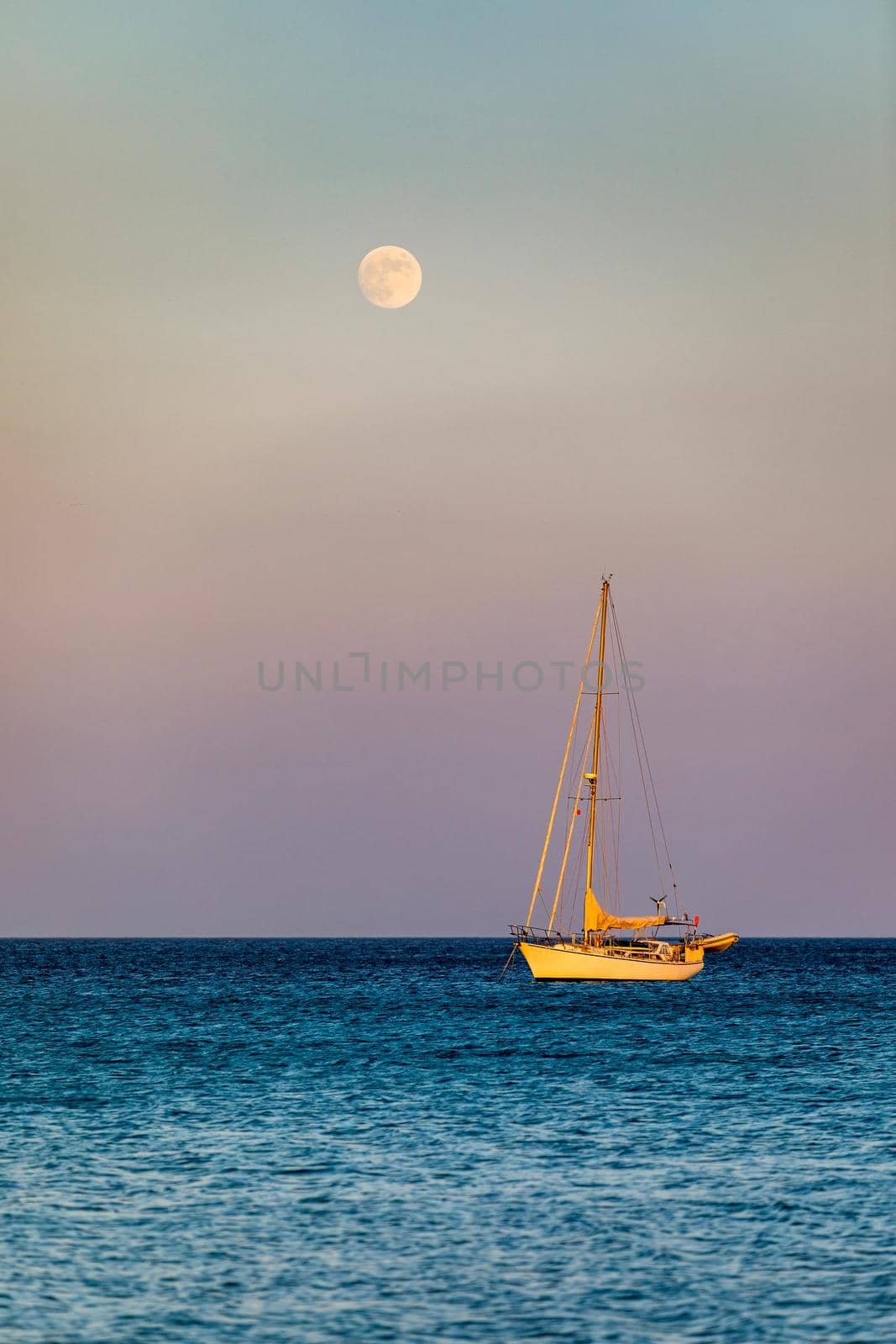 Full moon rising over the water with a small sailing boat in the foreground. Sailing boat with raising moon at sunset. Moon rising over the sea and yacht floating on the water surface. Sardinia, Italy