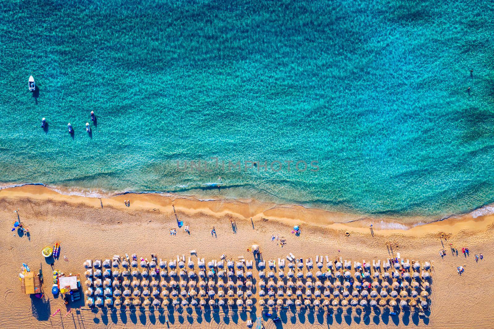 Aerial view of sandy beach with colorful umbrellas, swimming people in sea bay with transparent blue water at sunset in summer. Aerial top view on the beach, umbrellas, sand and sea waves.
