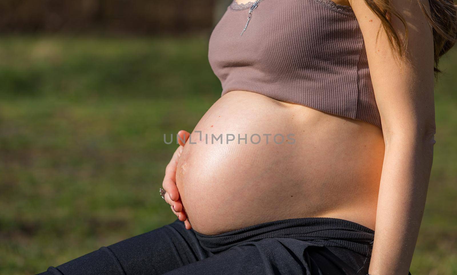 Beautiful pregnant woman feeling happy in nature while taking care of her child. Young expecting mother holding baby in pregnant belly. Maternity prenatal care and woman pregnancy concept.