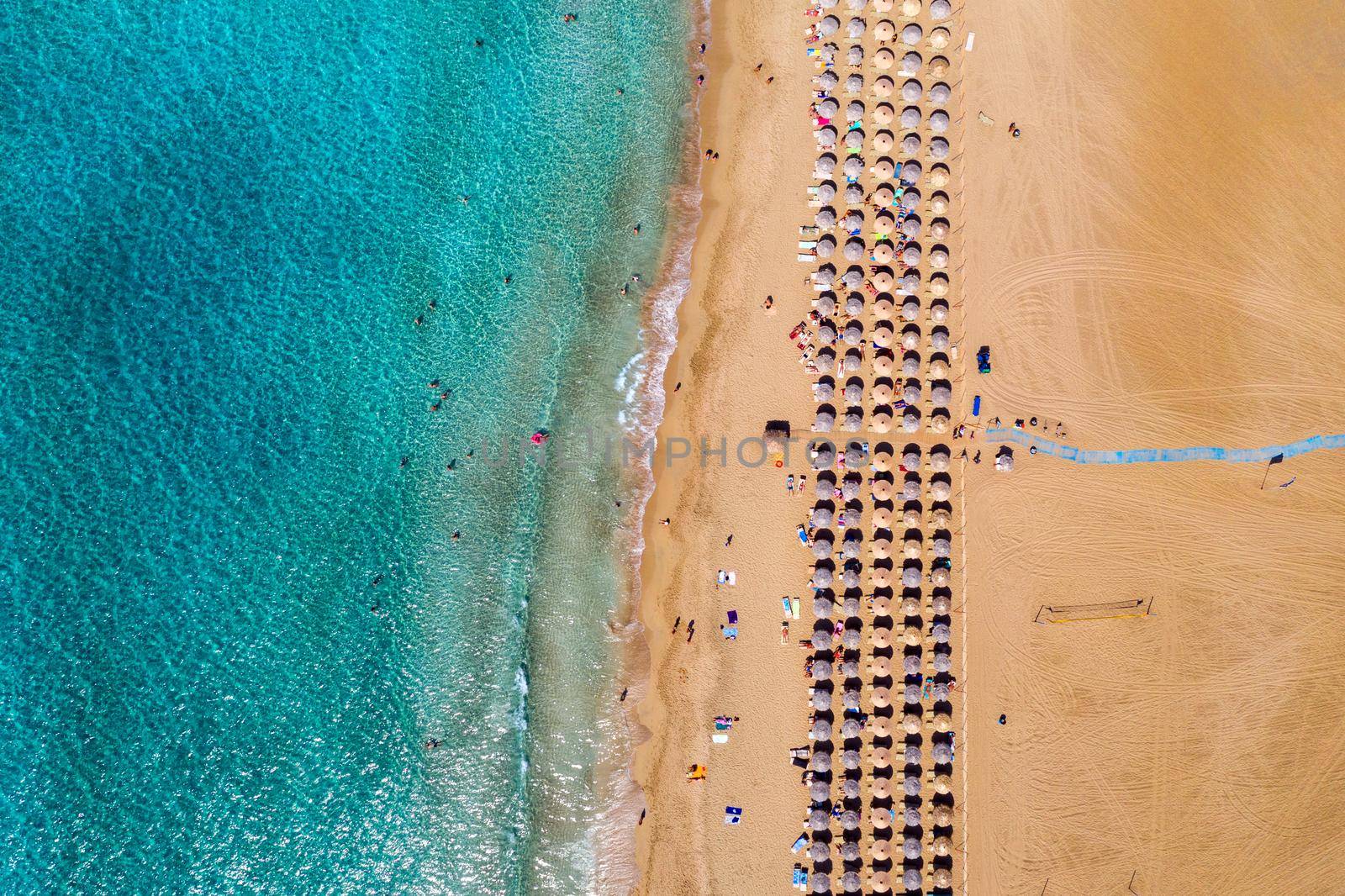 Aerial view of sandy beach with colorful umbrellas, swimming people in sea bay with transparent blue water at sunset in summer. Aerial top view on the beach, umbrellas, sand and sea waves.