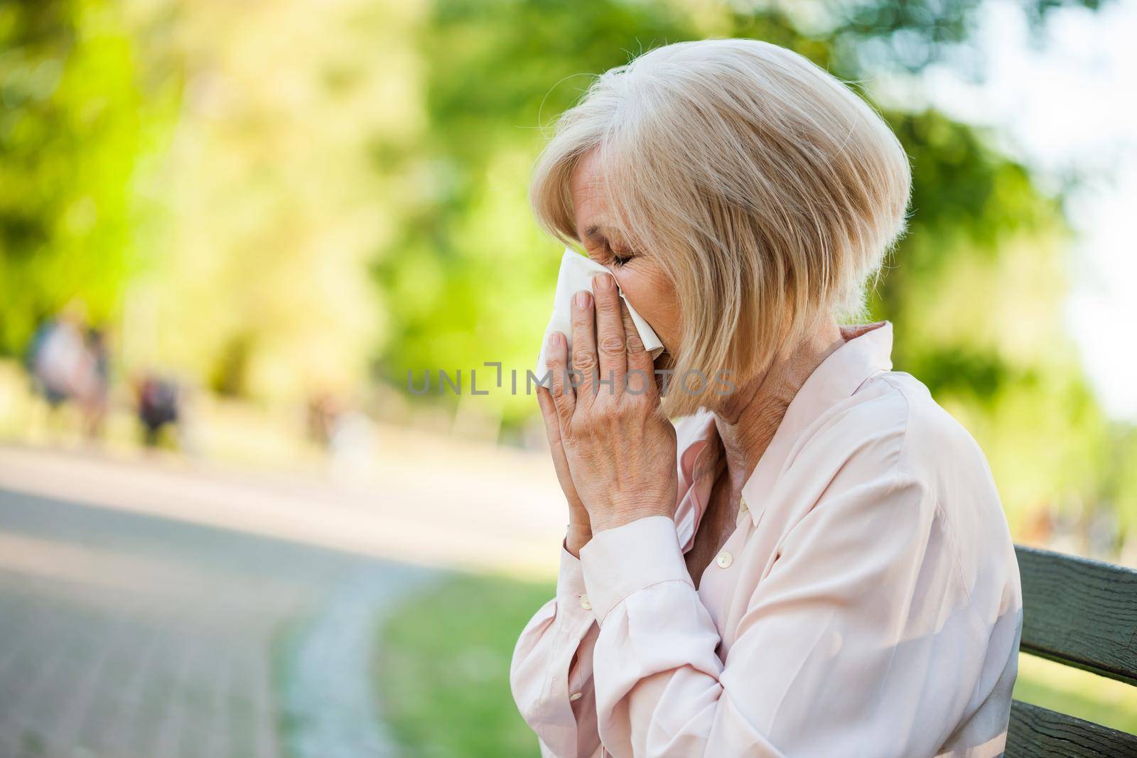 Adult woman is sitting in park and blowing nose. She is having allergy.