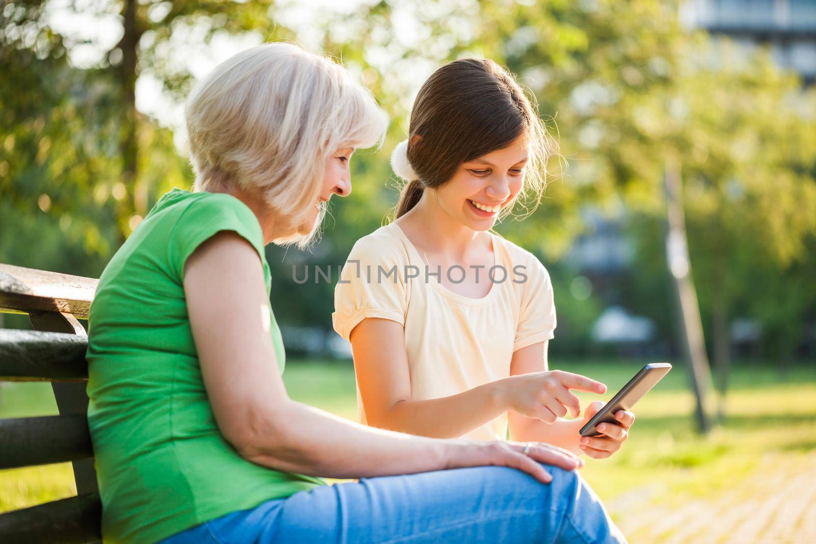 Grandmother and granddaughter are sitting in park and using mobile phone.