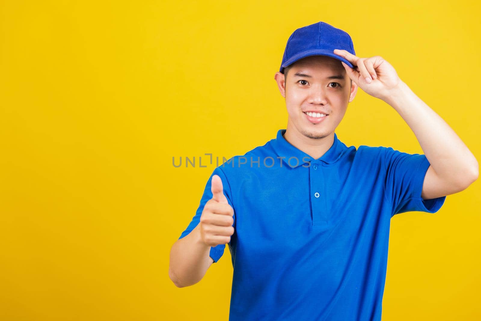 Portrait professional attractive delivery happy man standing he smile wearing blue t-shirt and cap uniform showing thumb up gesture looking to camera, studio shot isolated on yellow background