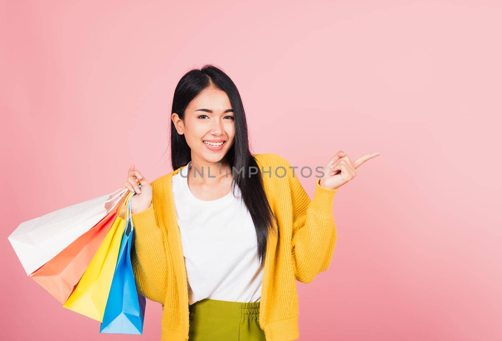 Portrait of Asian happy beautiful young woman shopper smiling standing excited holding online shopping bags multicolor in summer pointing finger to copy space, studio shot isolated on pink background