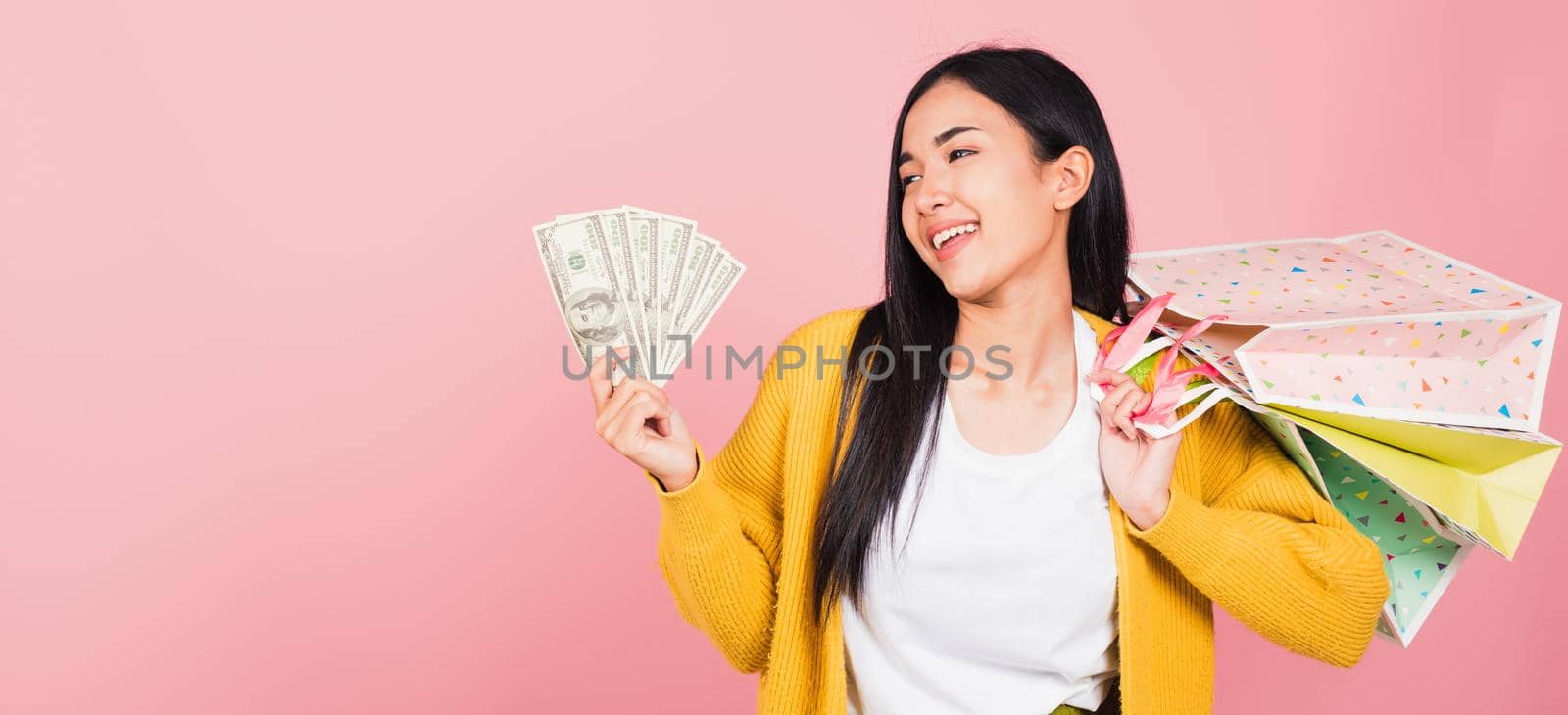 Portrait Asian happy beautiful young woman shopper smile excited holding online shopping bags colorful multicolor and dollar money banknotes on hand in summer, studio shot isolated on pink background
