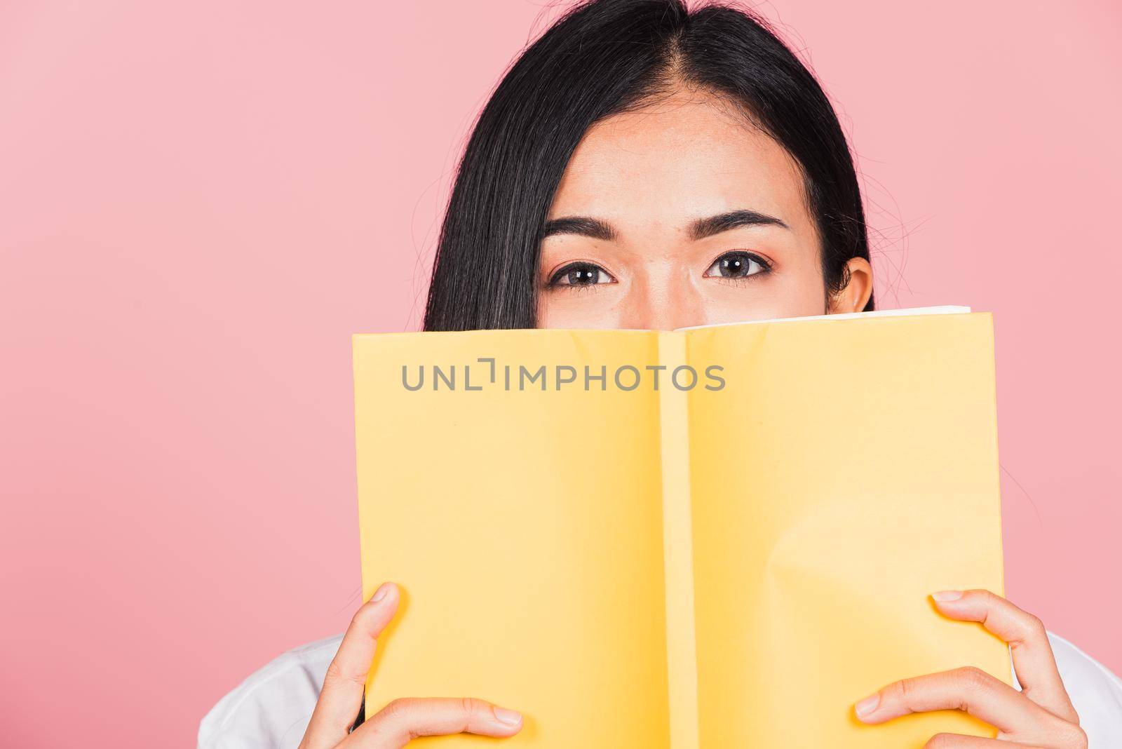 Portrait of beautiful Asian young woman teen smile covering her face with yellow book, female person hiding behind an open book show only eyes, studio shot isolated on pink background