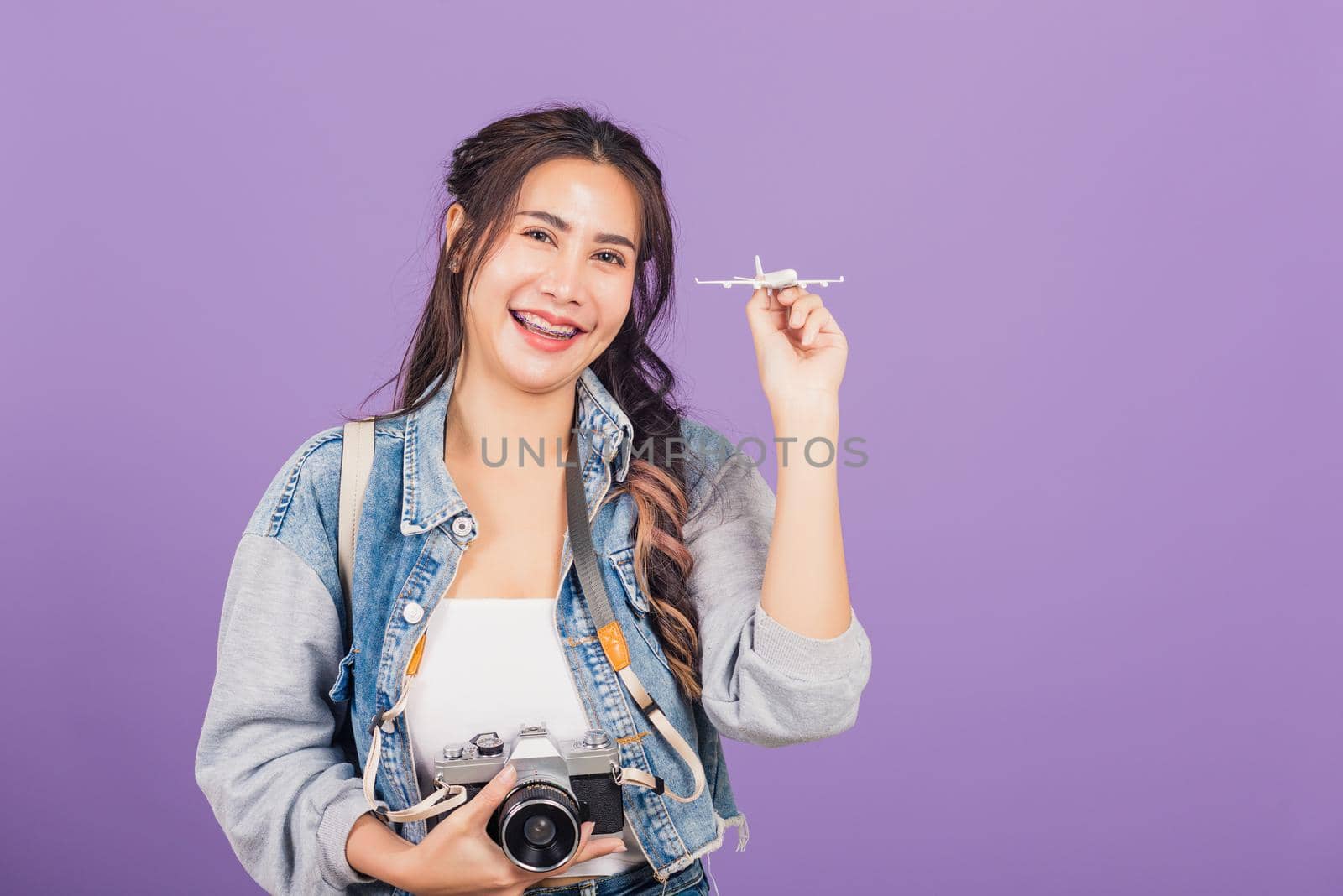 Happy Asian portrait beautiful young woman excited smiling holding airplane toy and retro vintage photo camera, Thai female ready travel trip isolated on purple background, tourism and vacation