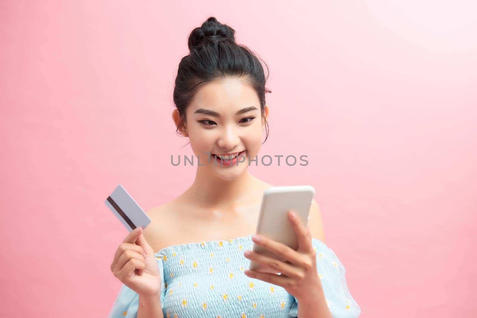 Portrait of a happy young woman using mobile phone, holding plastic credit card isolated over pink background