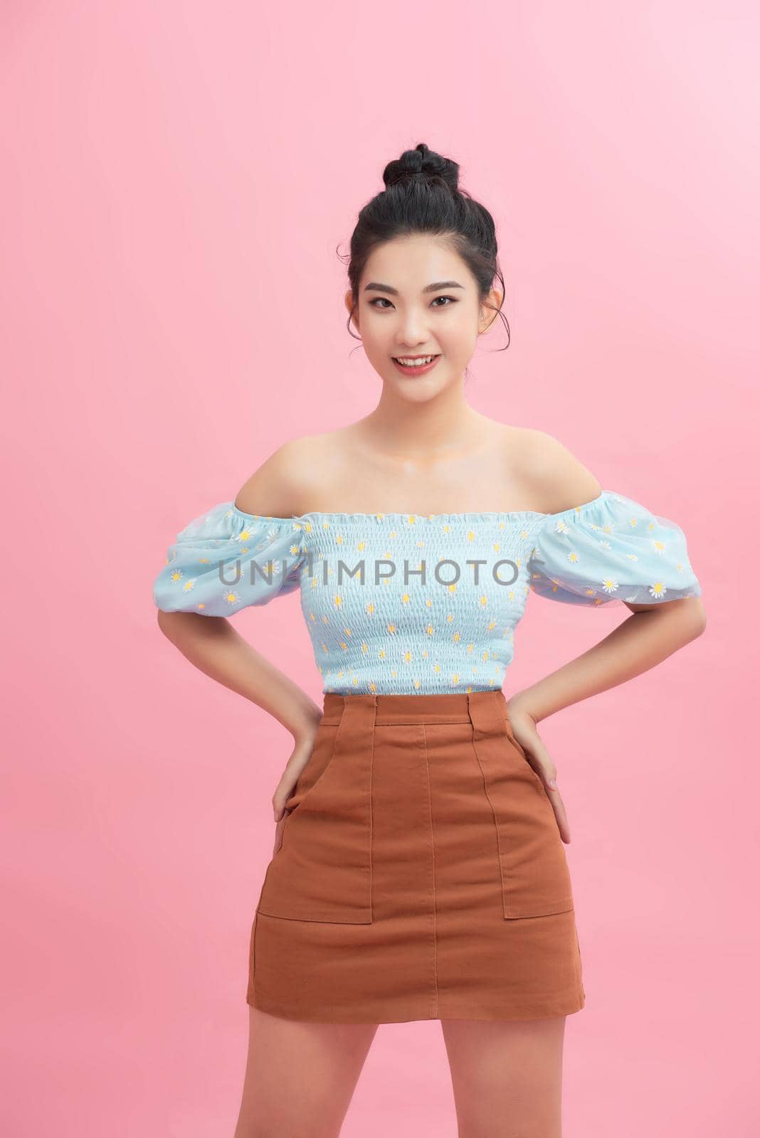  Smiling young woman standing with arms akimbo on waist isolated on pink pastel wall  by makidotvn