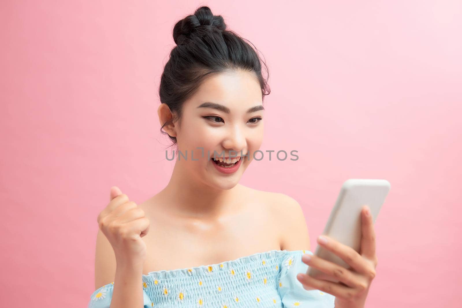 Close-up portrait of beautiful screaming young girl holding smartphone showing winner gesture, isolated on pink background