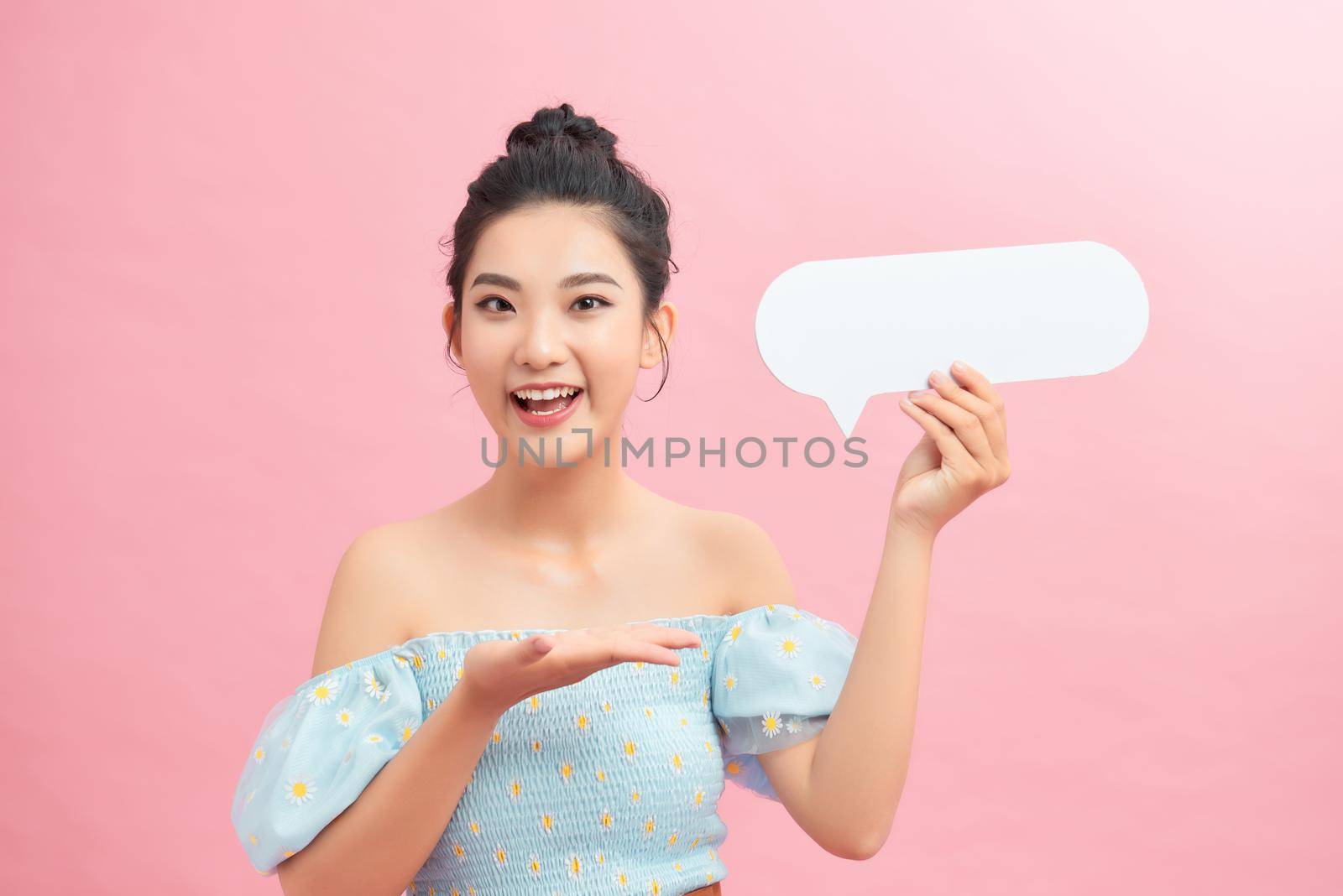 Surprised happy woman holding blank speech bubble and looking at the camera over pink background