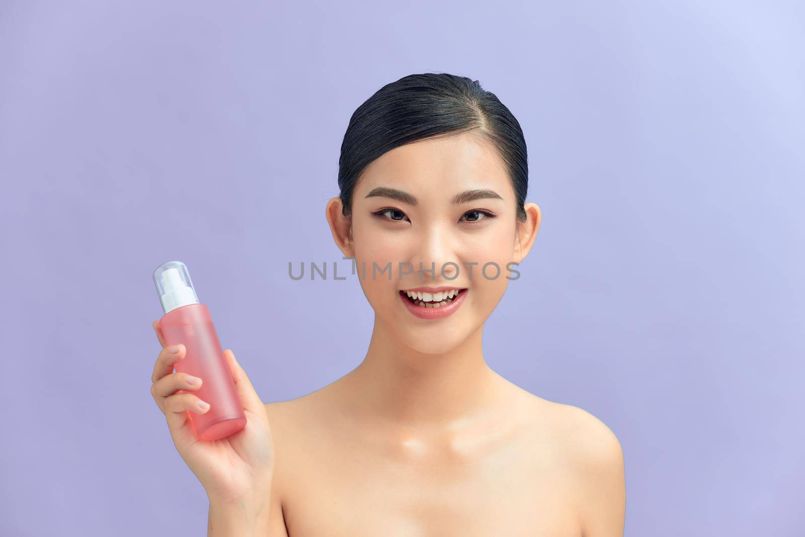 Beauty portrait of pretty young topless woman showing cosmetic skin oil in bottle isolated over purple background