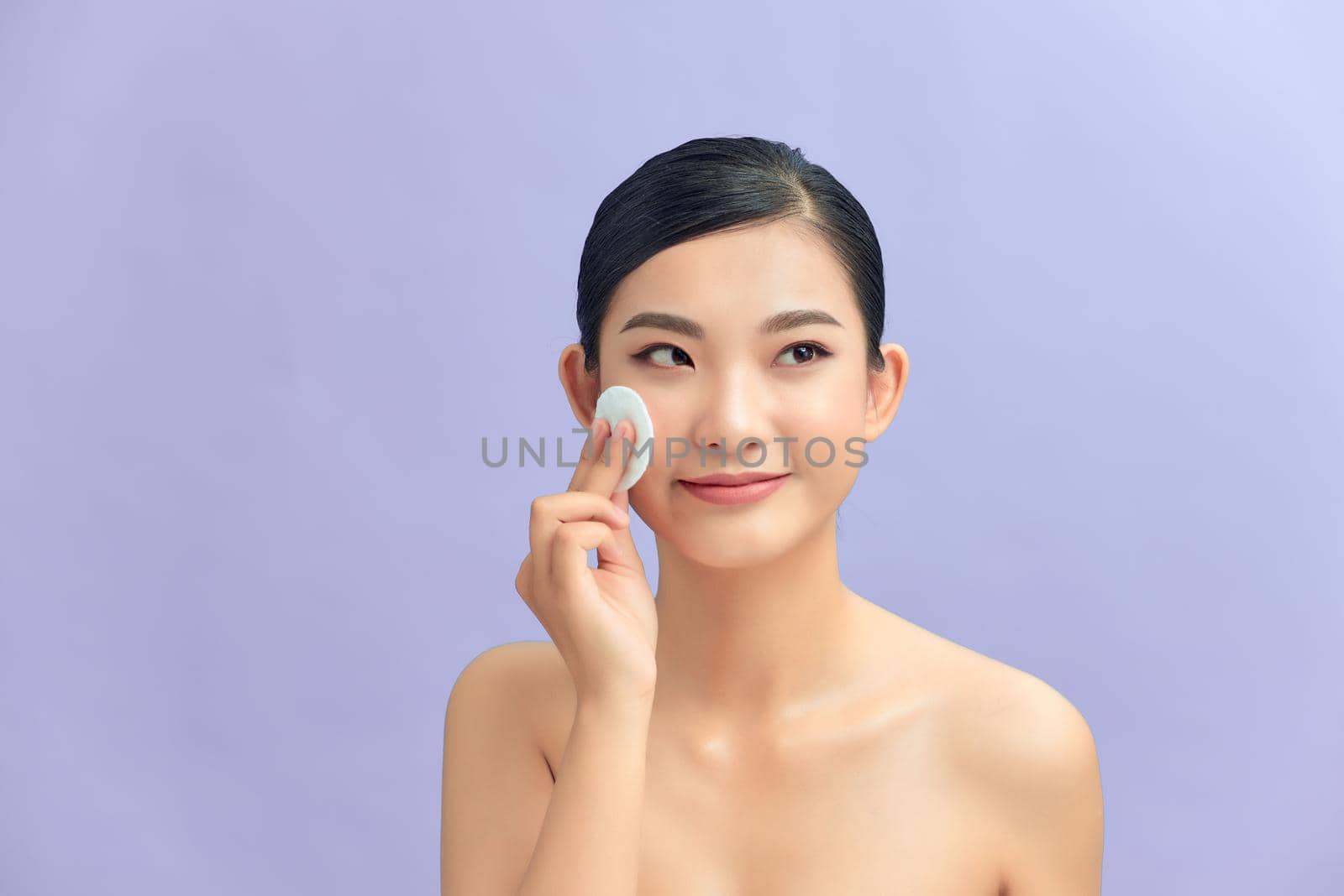 Crop attractive young female cleansing face with a cotton pad on a purple background in studio