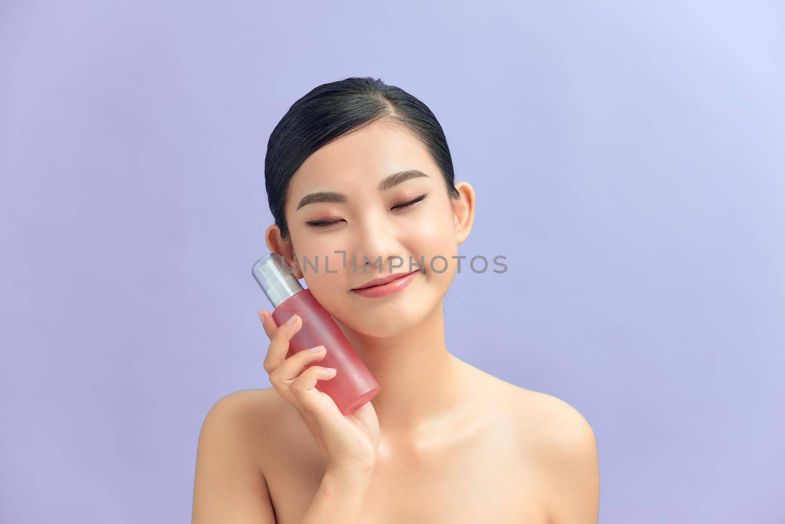 Beauty portrait of a pretty young topless woman wearing makeup isolated over purple background, showing cosmetic skin oil in a bottle by makidotvn