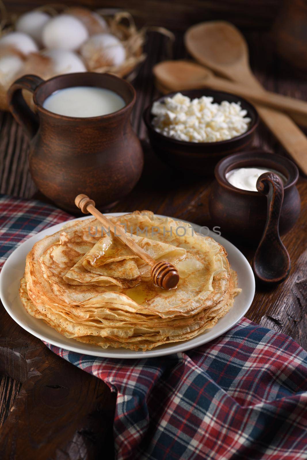 Homemade thin pancakes with honey stacked in a stack, on a wooden table with a mug of milk, a pot of sour cream and eggs in a basket. Traditional Slavonian, pagan holiday (Maslinitsa). Country style food.