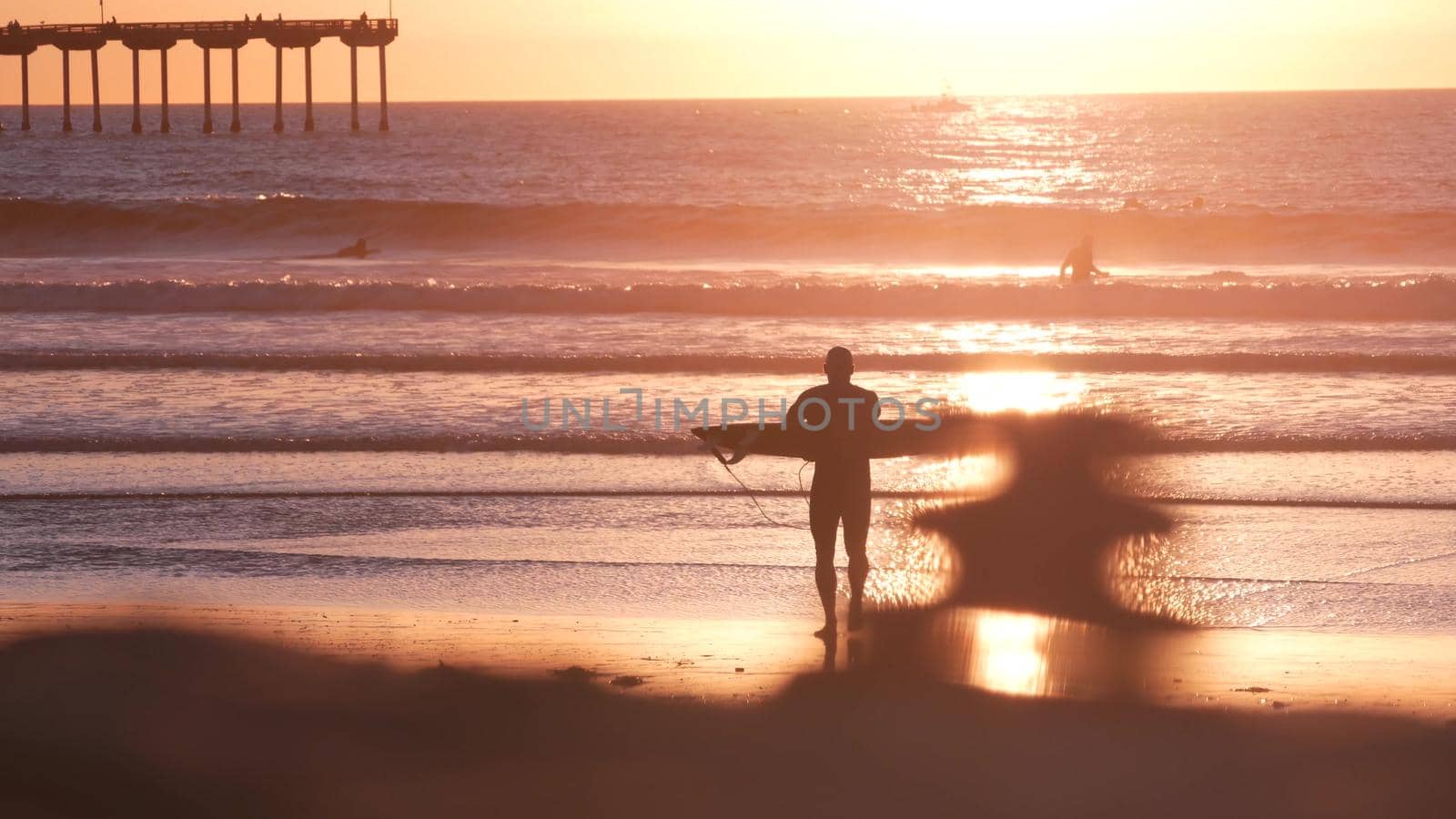 San Diego, California USA - 28 Nov 2020: People surfing by Ocean Beach pier on piles at sunset. Surfers on surfboards in water waves. Vacations on pacific coast or shore. Recreational sport hobby.