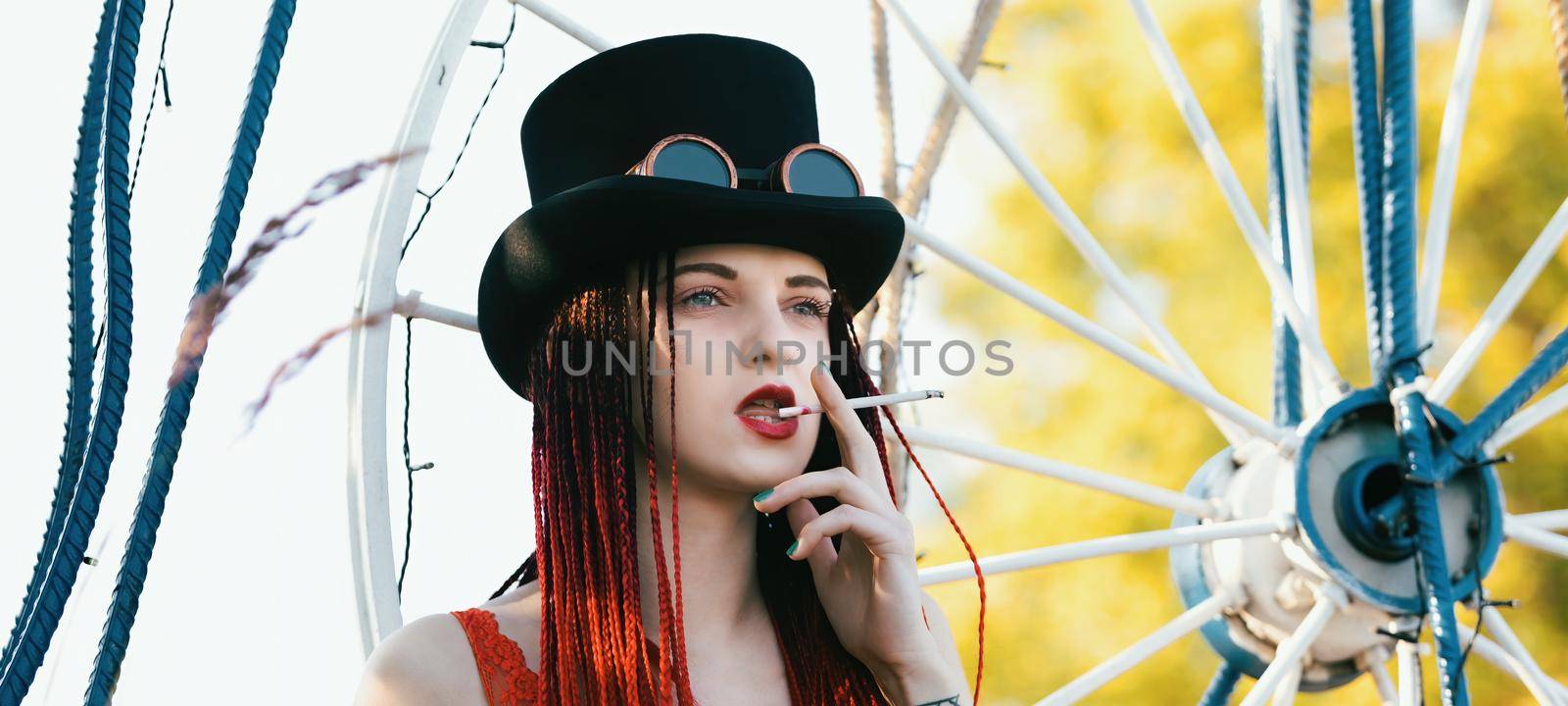 Glamorous girl with scarlet dreadlocks, red swimsuit, black hat and welding glasses posing outdoor with a cigarette 