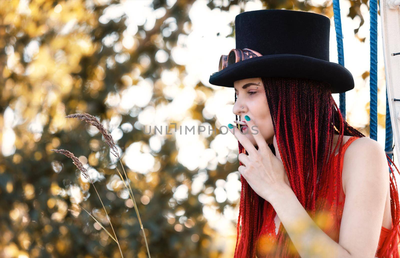 Glamorous girl with scarlet dreadlocks, red swimsuit, black hat and welding glasses posing outdoor with a cigarette 