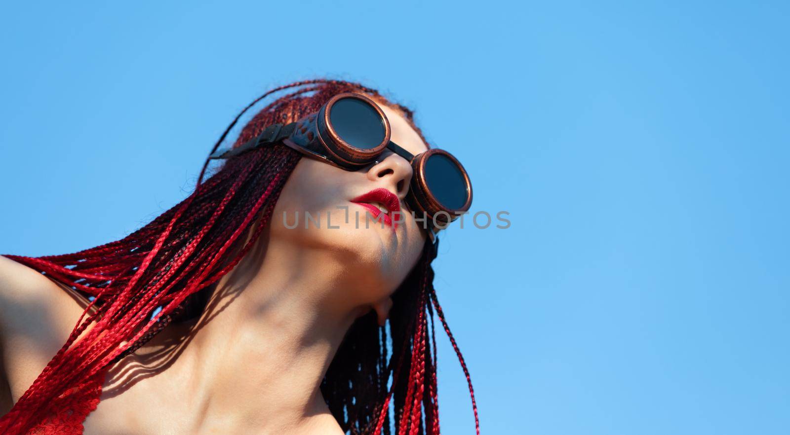 Glamorous girl with scarlet dreadlocks, a red swimsuit and welding glasses poses on the blue sky background. Free space for your text