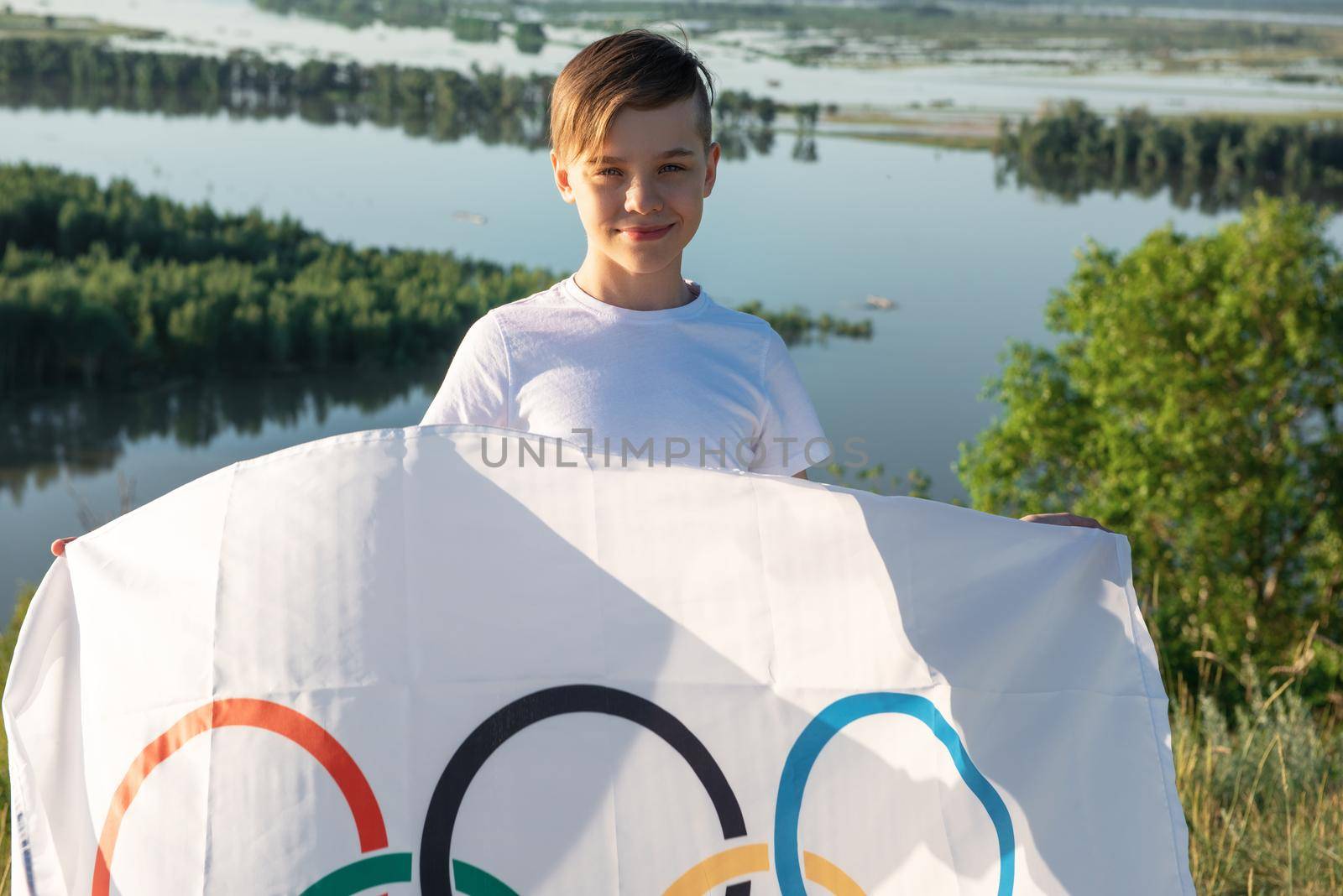 Portrait of boy waving flag the Olympic Games outdoors over cloudy sunset sky. Children sports fan. Summer olympic games concept. 08.06.2021, Barnaul Russia.
