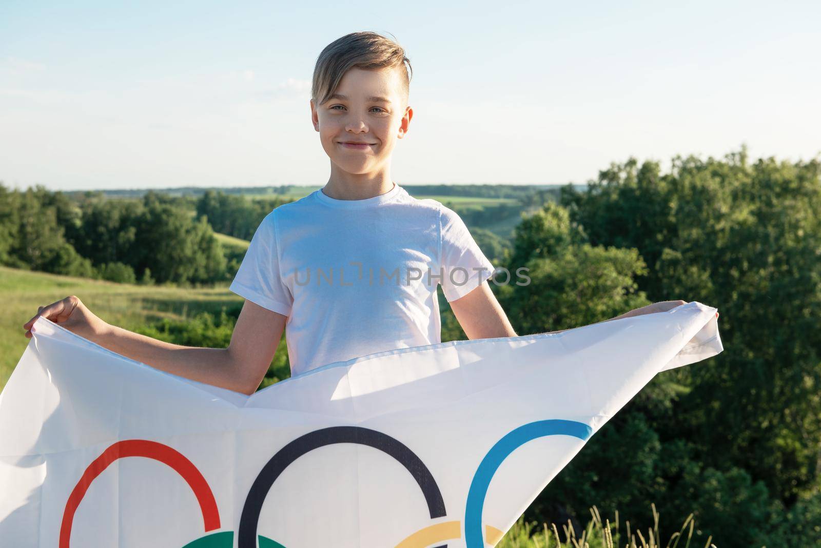 Blonde boy waving waving flag the Olympic Games outdoors over blue sky at the river bank by rusak