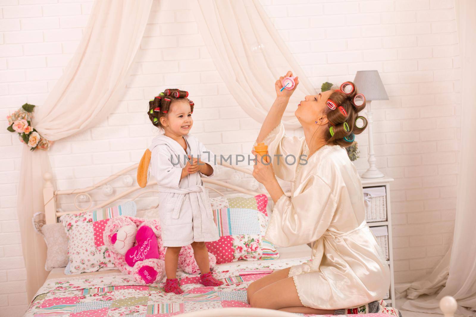 A charming little girl with her mother in hair curlers play with soap balls. Women's day. The girl has fun with her mom by Try_my_best