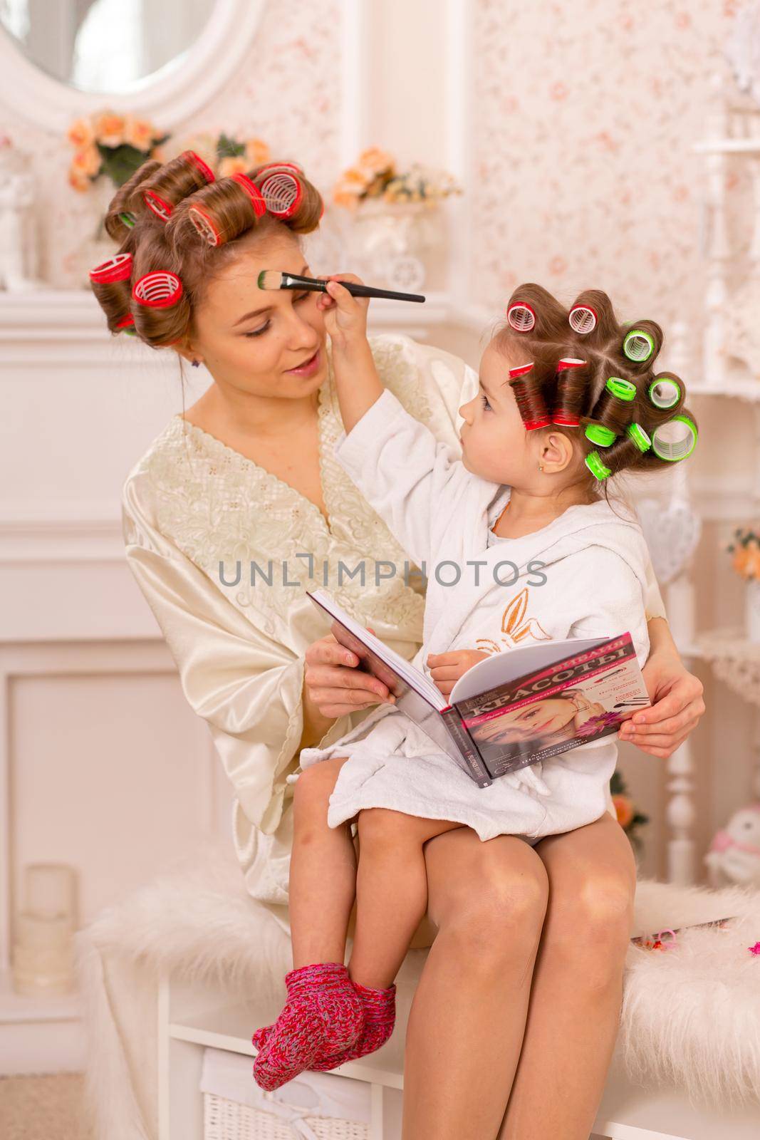 Adorable little girl with her mother in hair curlers apply makeup. Mom teaches daughter to use cosmetics. Beauty day. Girls are such girls.