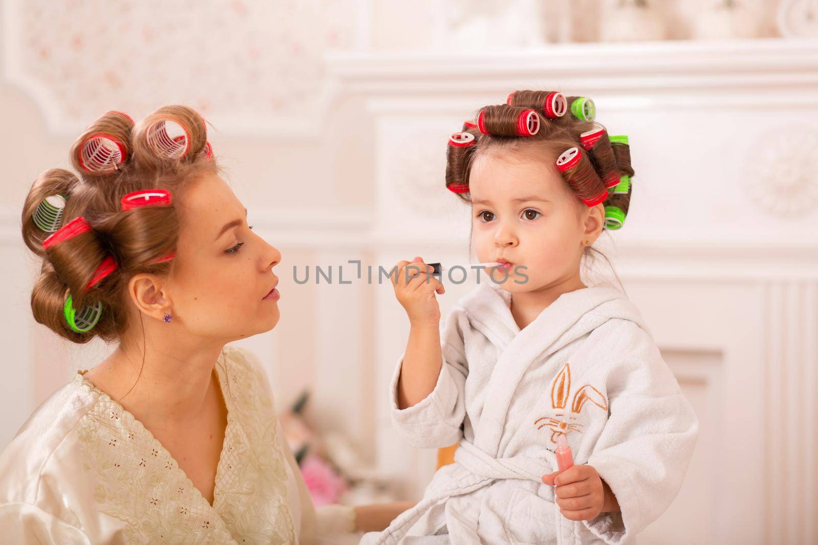 Adorable little girl with her mother in hair curlers apply makeup. Mom teaches daughter to use cosmetics. Beauty day. Girls are such girls.