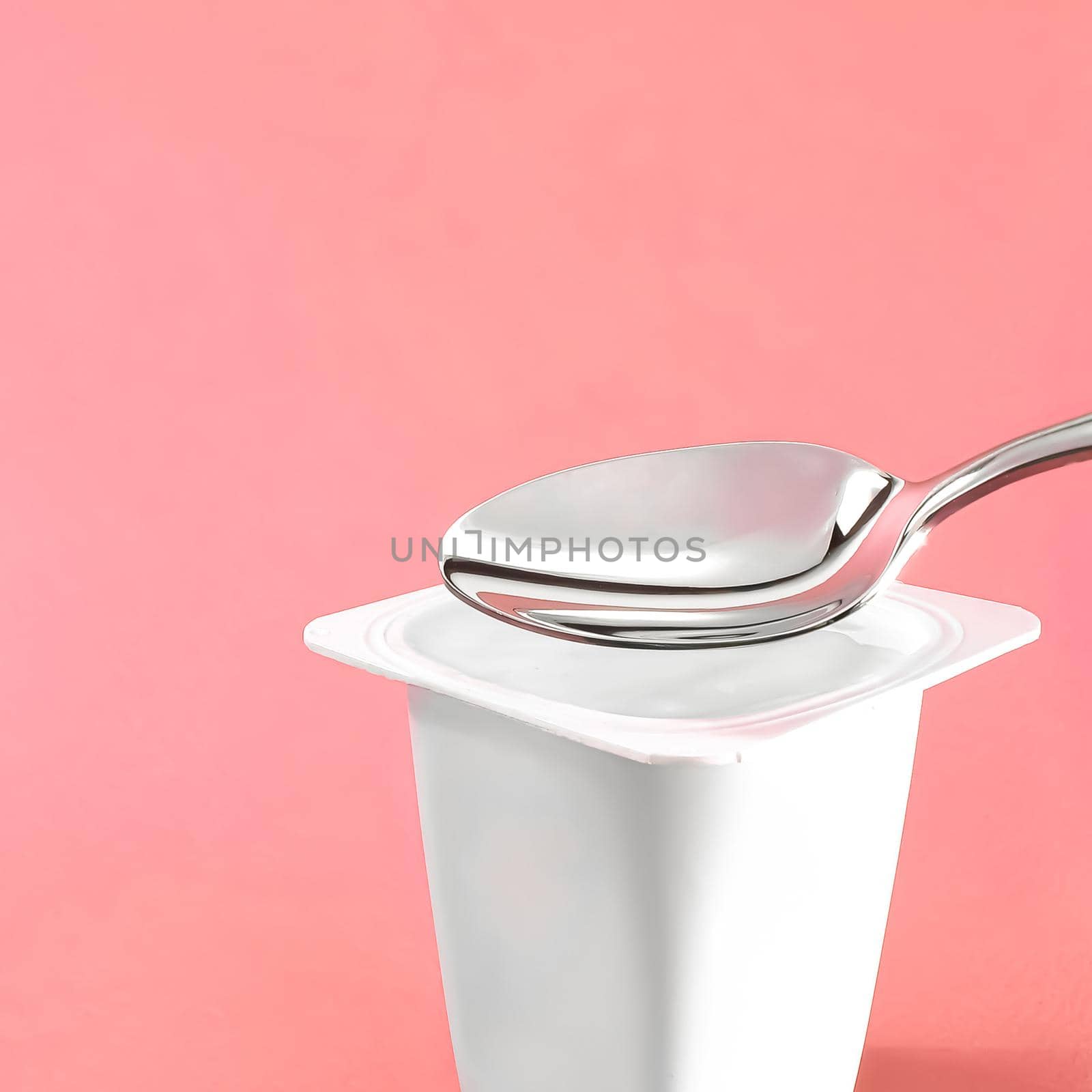Yogurt cup and silver spoon on pink background, white plastic container with yoghurt cream, fresh dairy product for healthy diet and nutrition balance by Anneleven