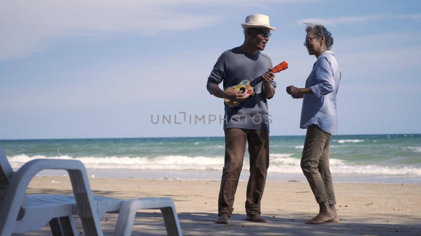 Happy senior couple relaxing outdoors singing and playing acoustic guitar at beach near sea sunny day, Mature man playing ukulele for his wife at sea, plan life insurance at retirement couple concept