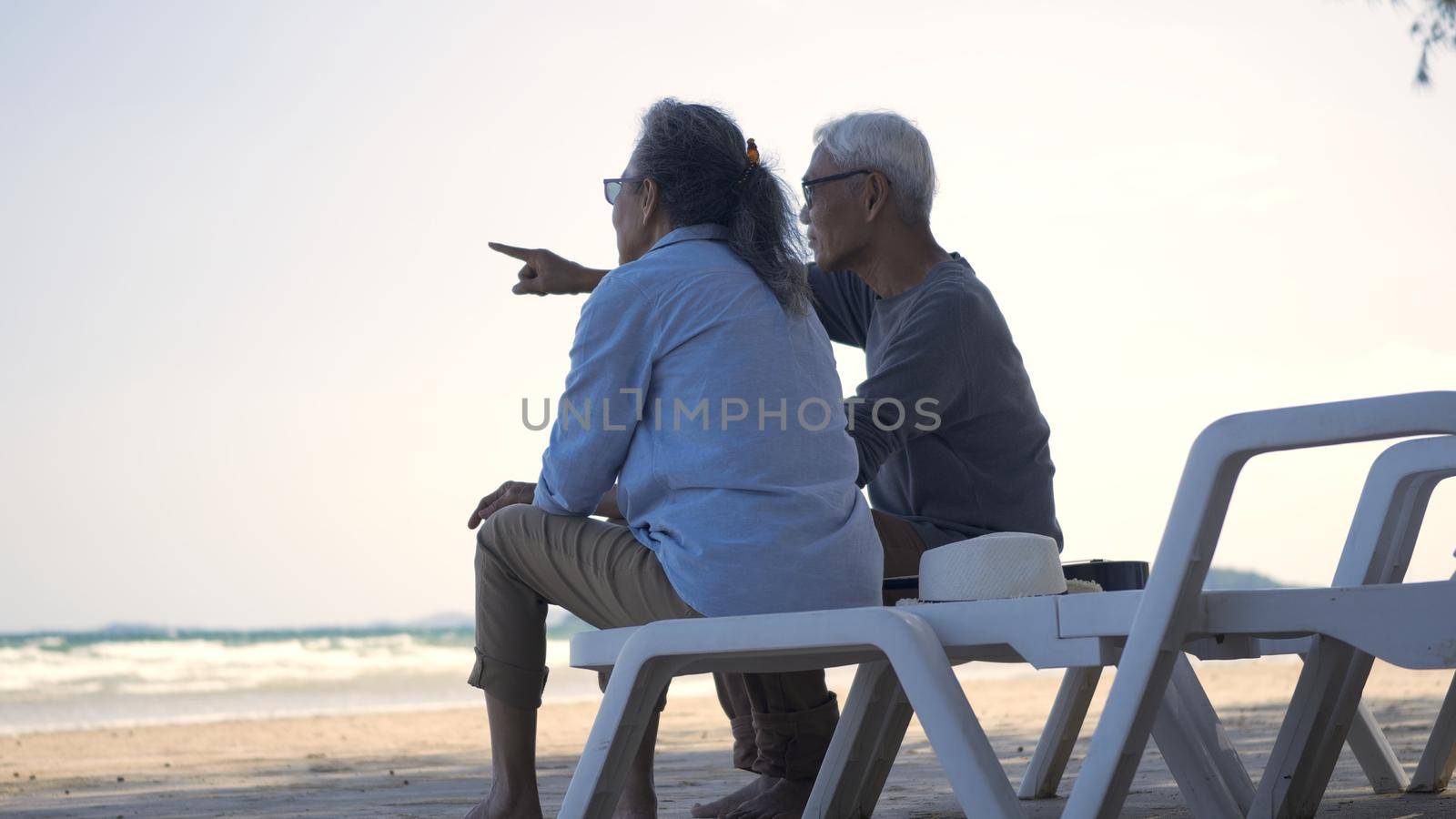 Happy Asian family, senior couple sitting on chairs with backs on beach travel vacation talking together, Romantic elderly enjoy Travel summer vacation, plan life insurance retirement couple concept