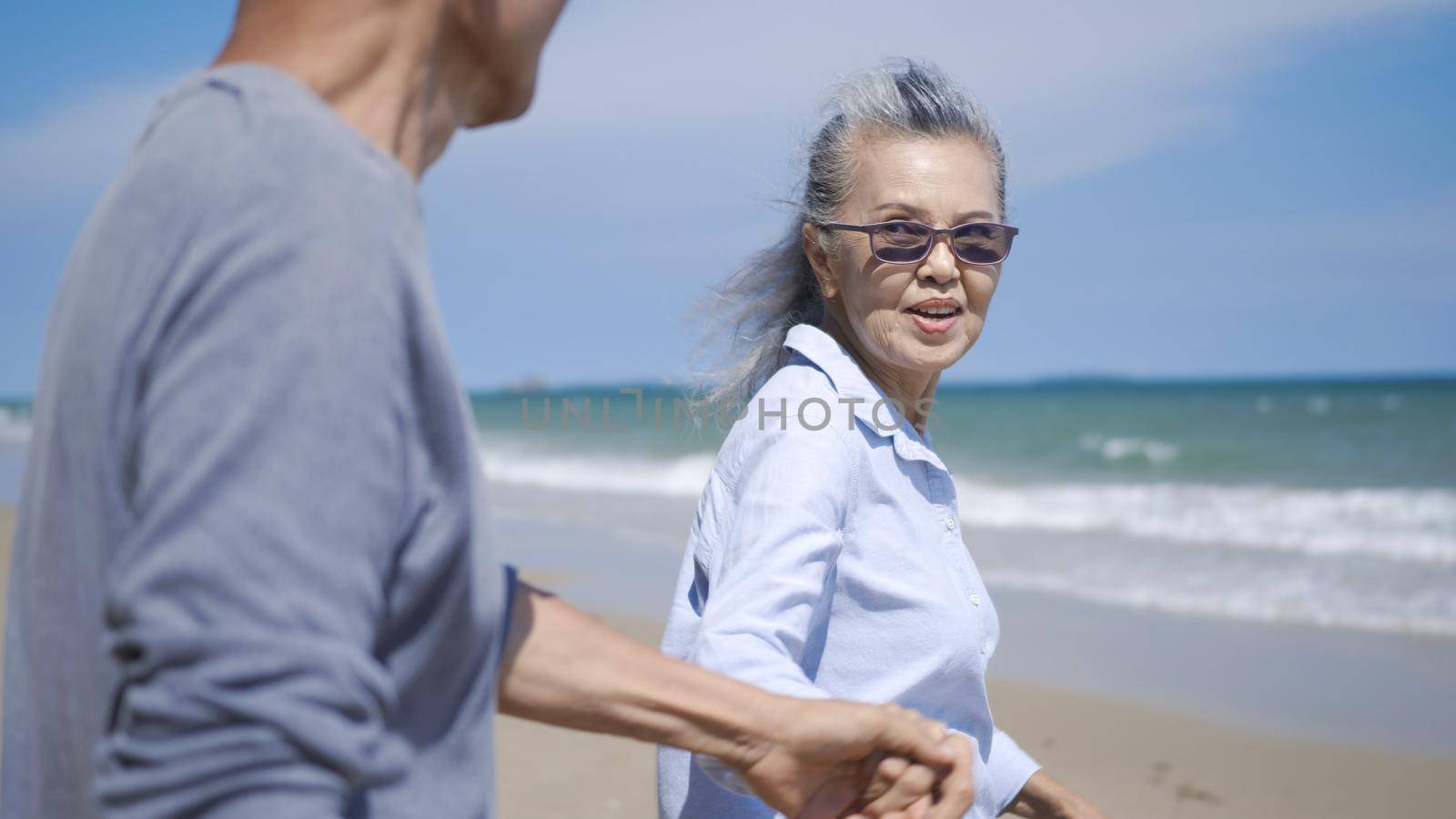 Happy Asian senior man and woman couple holding hands walking to the beach sunny with bright blue sky, Romantic elderly enjoy Travel summer vacation, plan life insurance at retirement couple concept