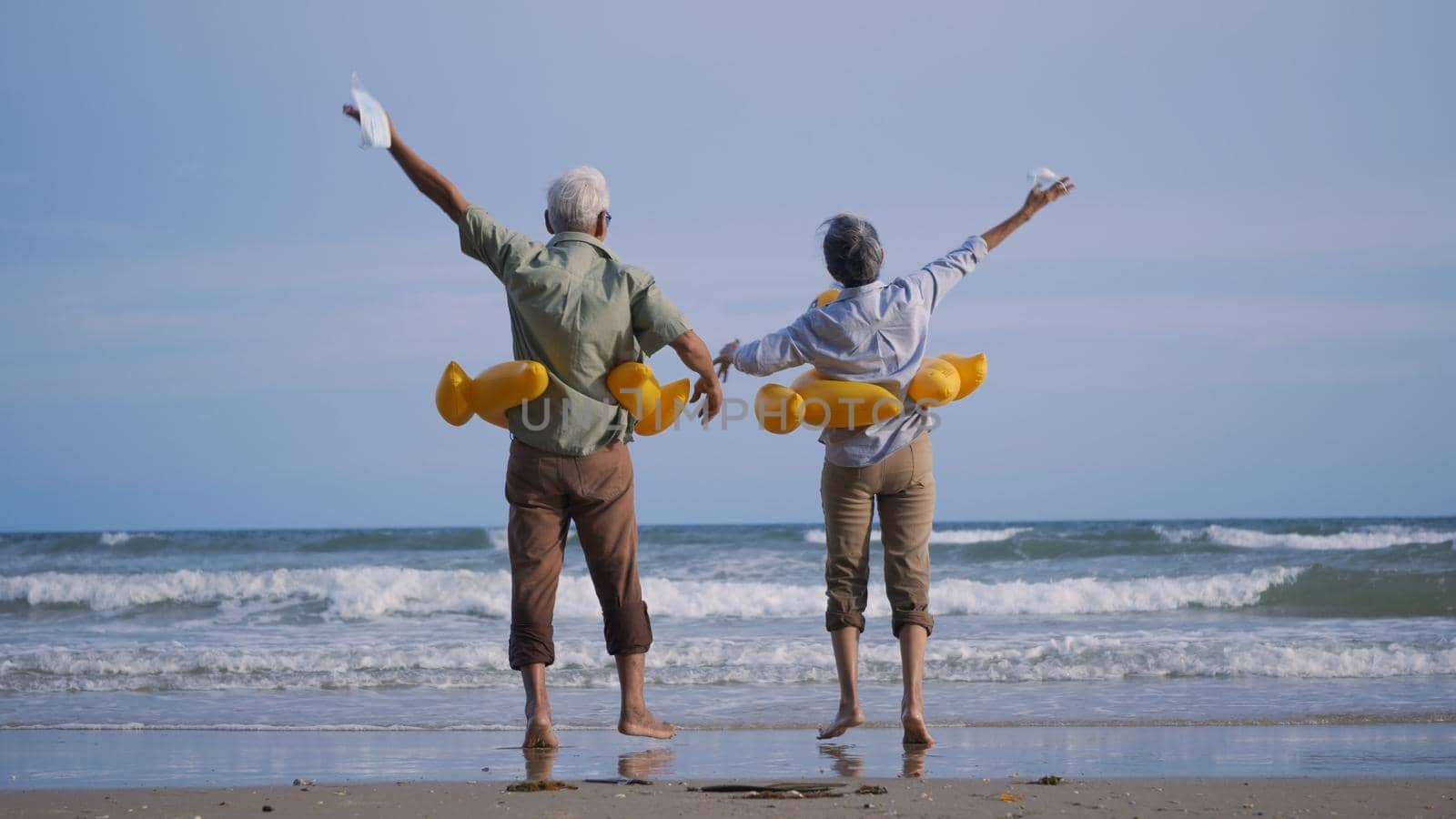 Happy senior couple wear rubber ring take off medical face mask and running to the beach enjoying life, relaxing after win coronavirus pandemic (COVID-19), plan life insurance at retirement couple