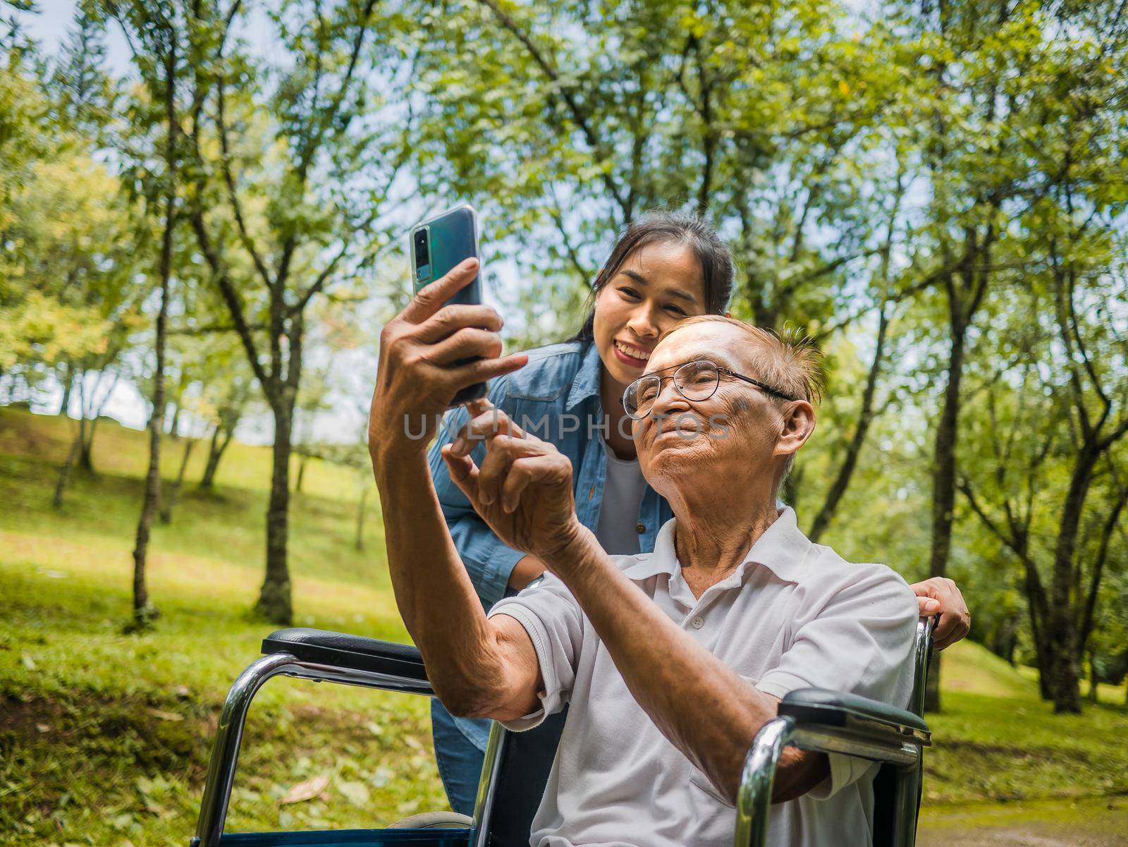 Grandfather in wheelchair and granddaughter talking video call with relatives via smartphone in park. Family life on vacation. by TEERASAK