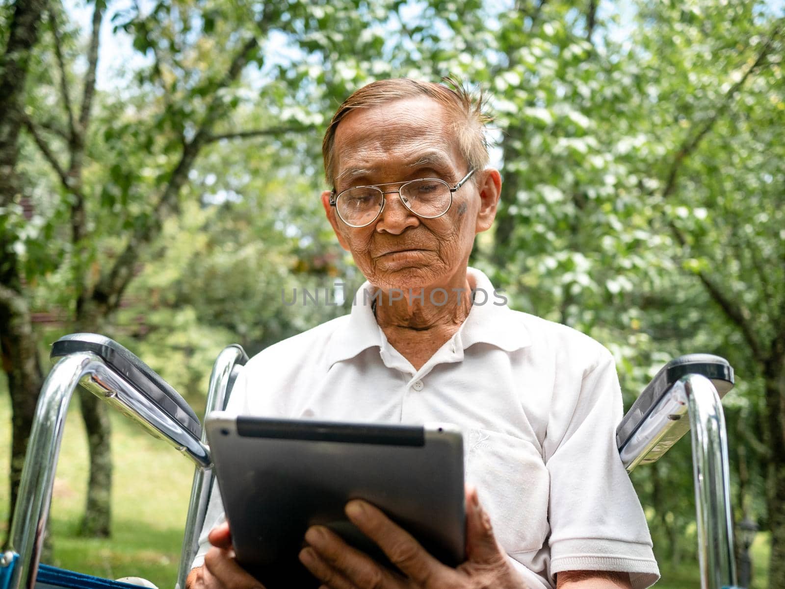 Senior man in wheelchair using digital tablet, looking at screen, reading e-book, chatting online with grandchild, watching movie in the park. by TEERASAK