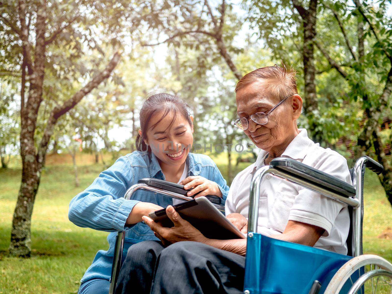 Grandfather in wheelchair and granddaughter having fun with digital tablet in the park. Family life on vacation. by TEERASAK