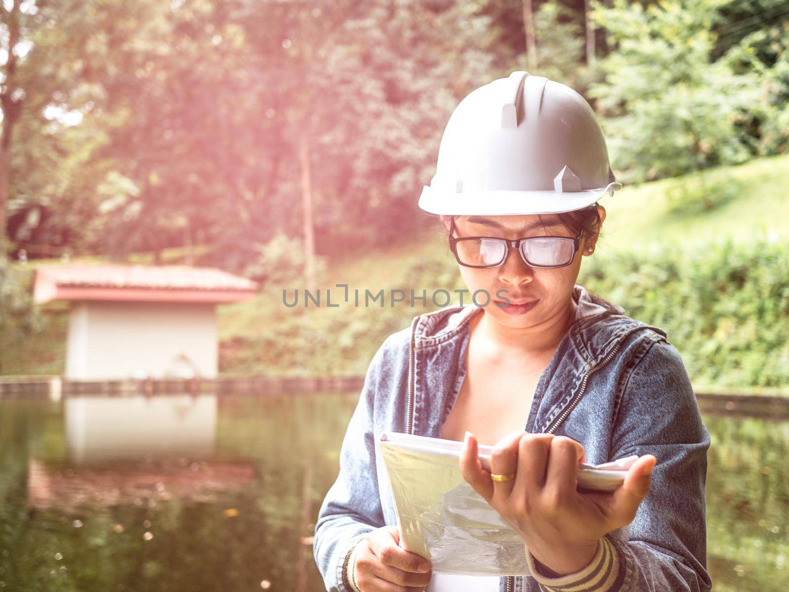 Female ecologist in safety hat working on site at the dam and stands reading the blueprints at the construction site.