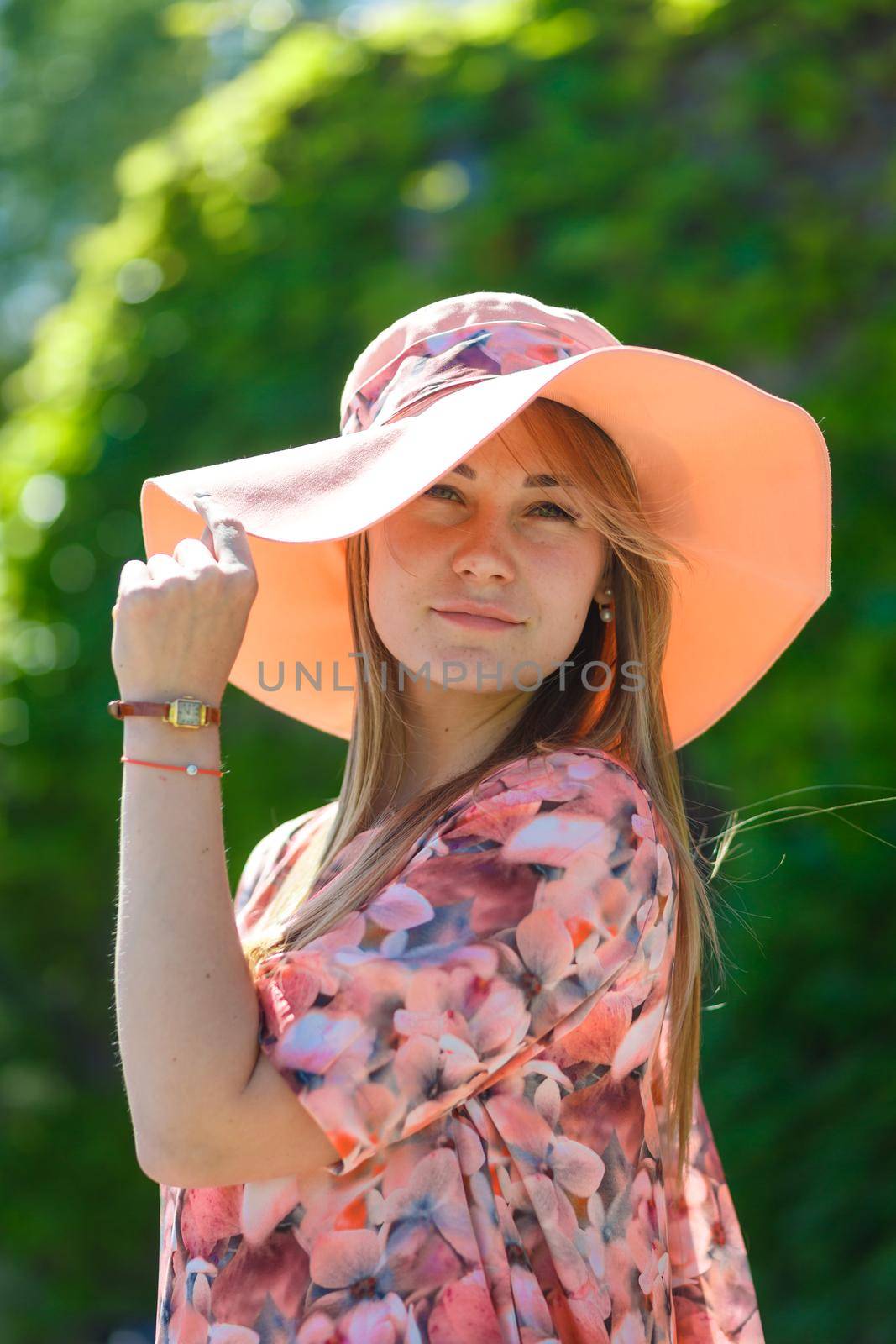 A charming girl in a light summer sundress and a pareo hat is walking in a green park. Enjoys warm sunny summer days.