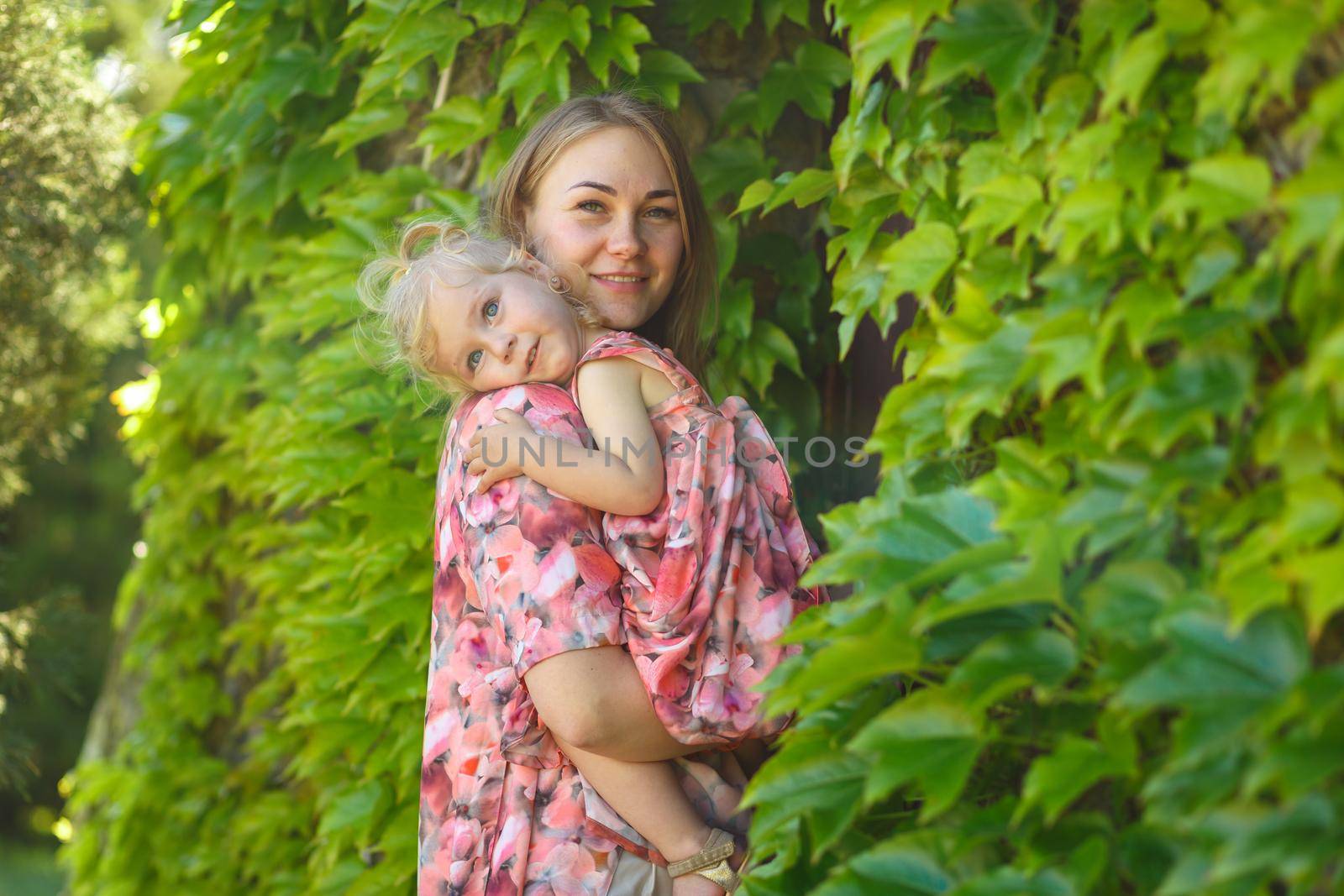 A charming girl in a light summer sundress walks in a green park with her little daughter, holding her in her arms. Enjoys warm sunny summer days.