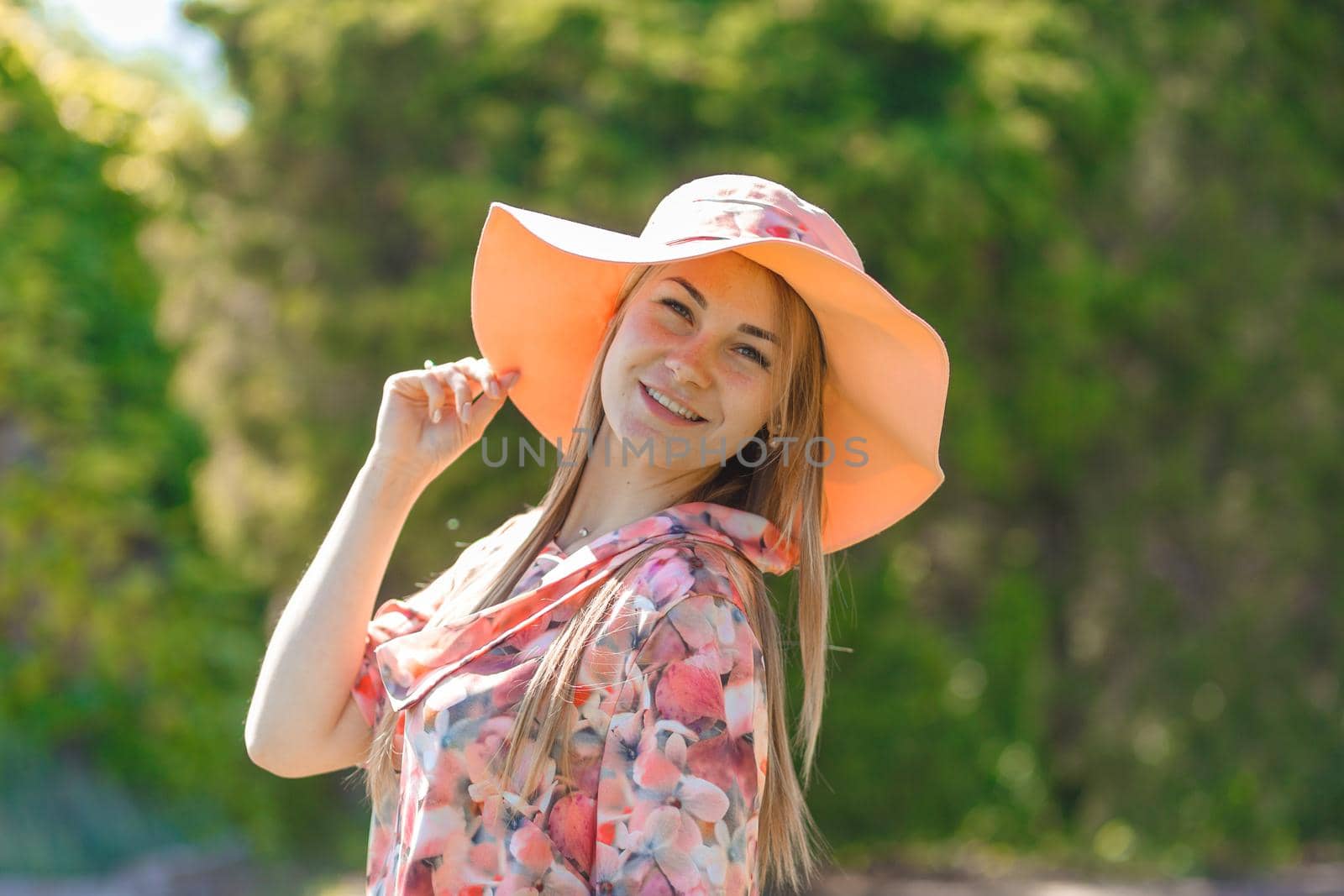A charming girl in a light summer sundress and a pareo hat is walking in a green park. Enjoys warm sunny summer days.