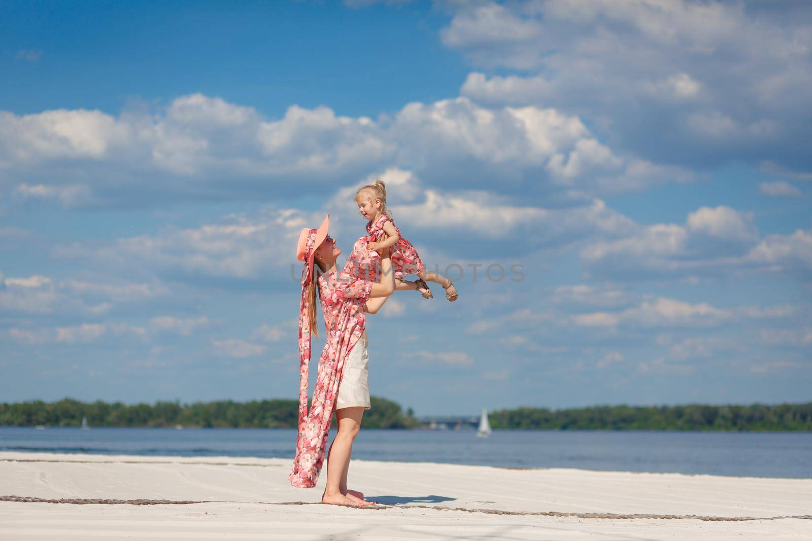 A charming girl in a light summer sundress walks on the sandy beach with her little daughter. Enjoys warm sunny summer days.