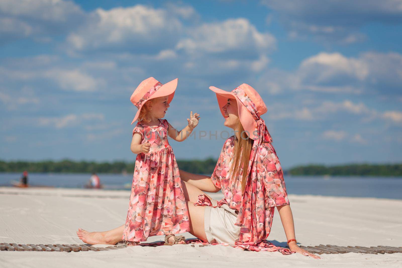 A little girl with her mother in matching beautiful sundresses plays in the sand on the beach. Stylish family look by Try_my_best