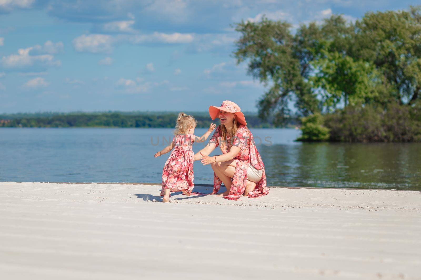 A charming girl in a light summer sundress walks on the sandy beach with her little daughter. Enjoys warm sunny summer days.