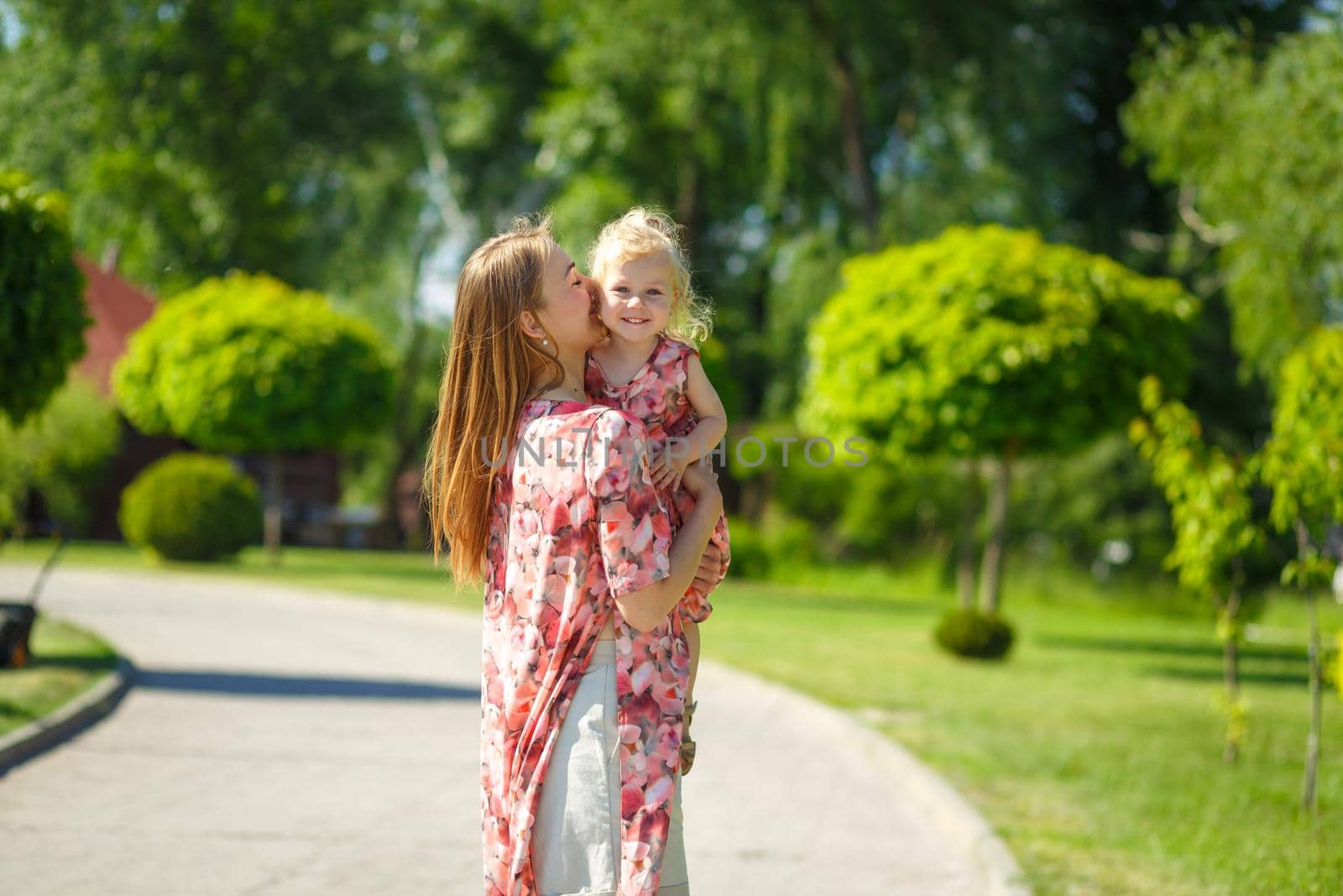A charming girl in a light summer sundress walks in a green park with her little daughter, holding her in her arms. Enjoys warm sunny summer days.
