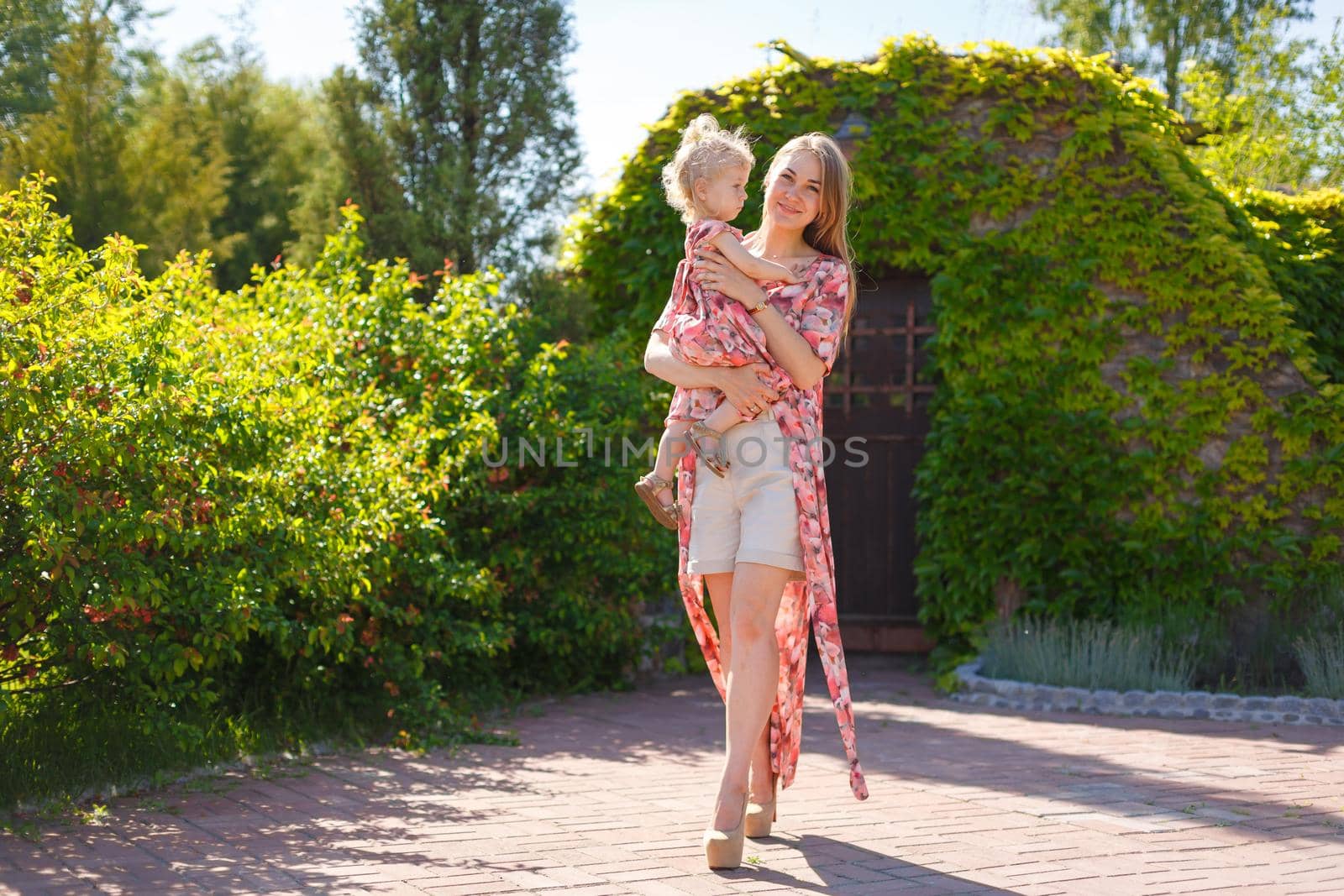 A charming girl in a light summer sundress walks in a green park with her little daughter, holding her in her arms. Enjoys warm sunny summer days.